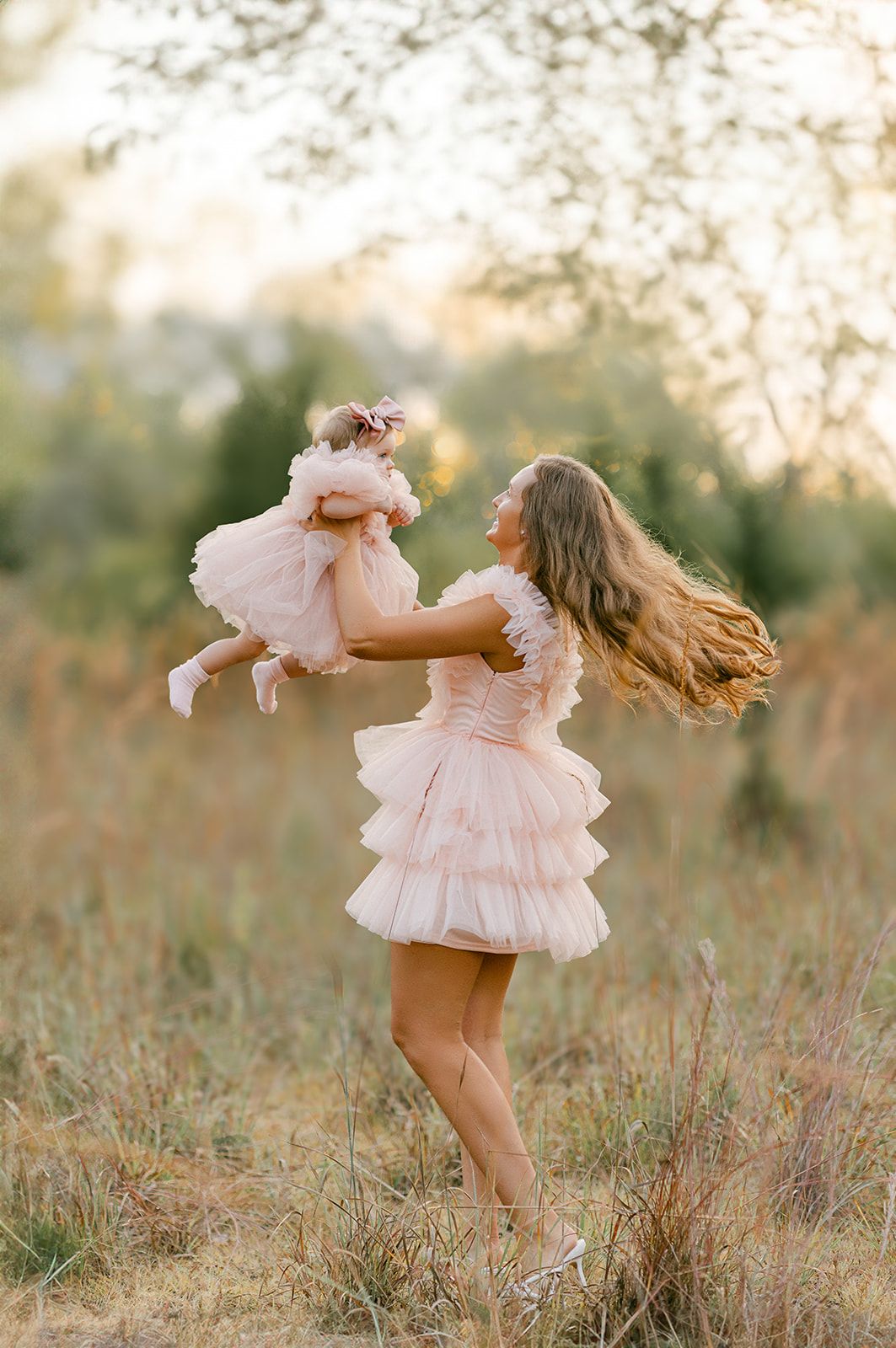 Mother in pink tiered dress lifts smiling baby girl in matching outfit in sunny meadow.