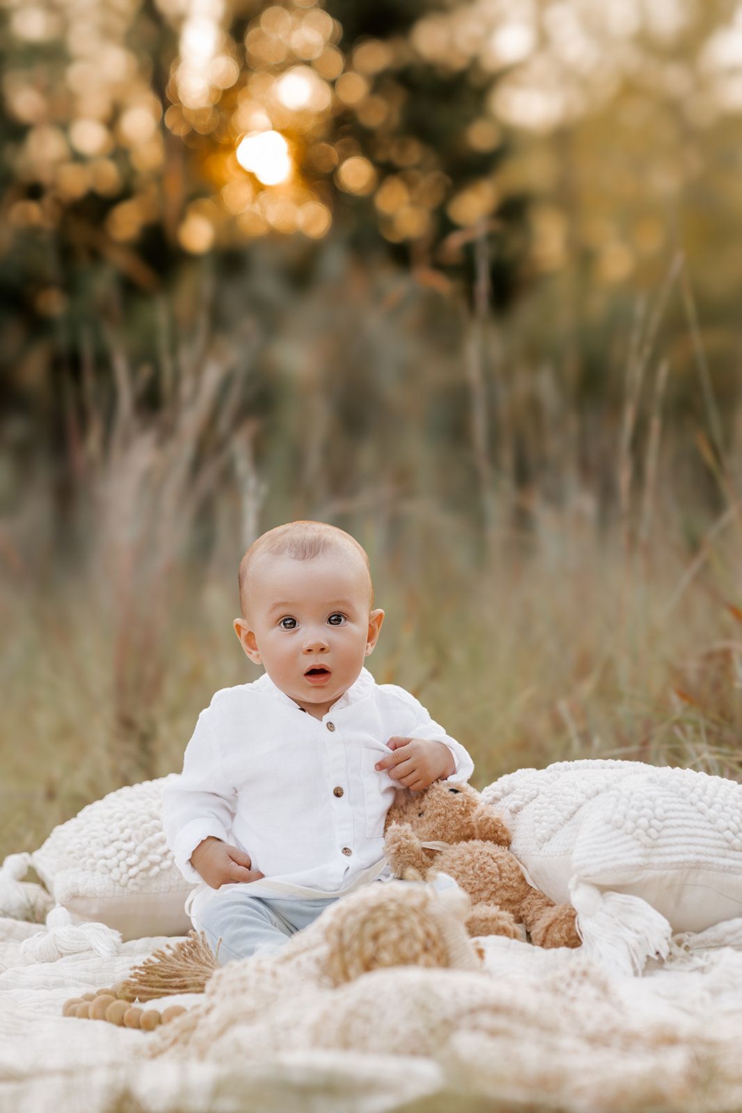 Adorable baby in white shirt sitting on blanket with teddy bear in golden sunset field.
