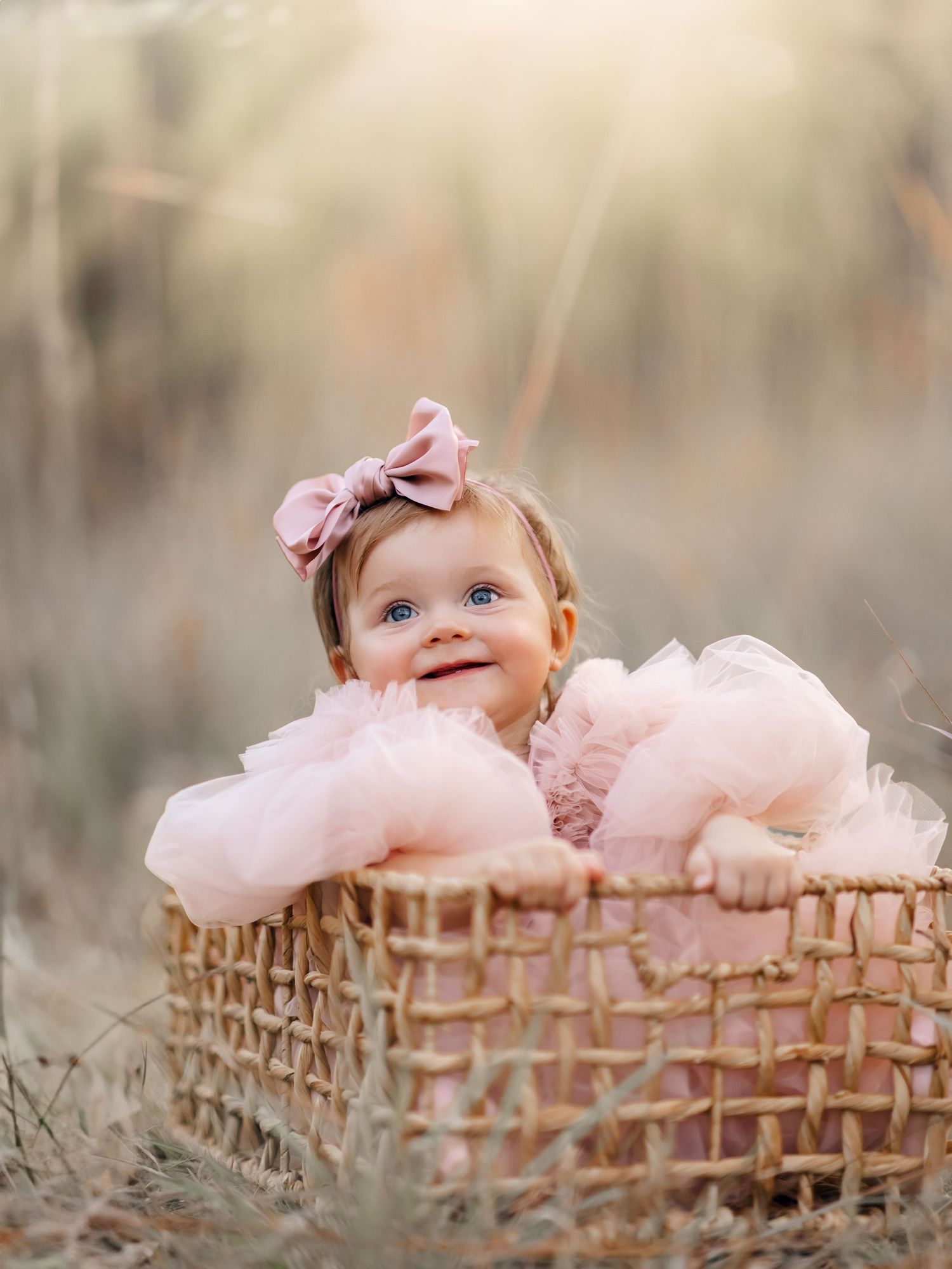 Adorable blue-eyed baby girl in pink tulle dress peeking over edge of wicker basket outdoors.