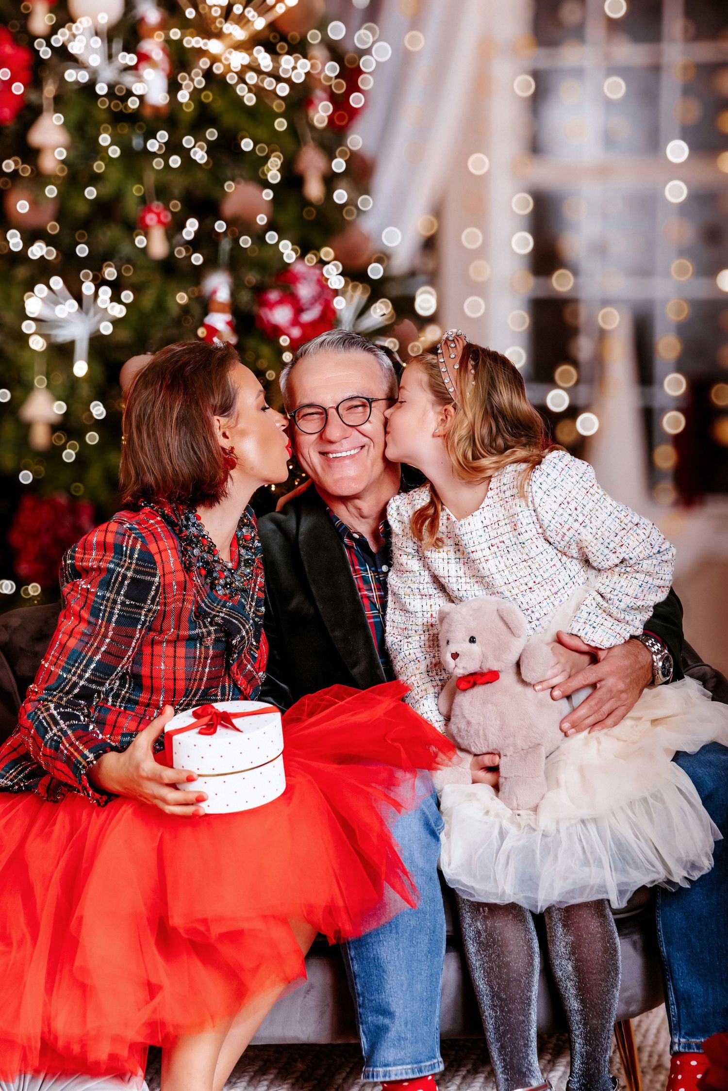 Grandparents receive cheek kisses from granddaughter holding teddy bear near Christmas tree.