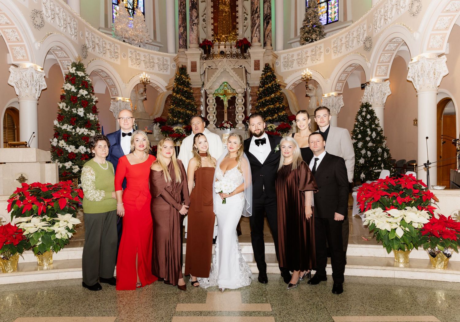 Wedding family portrait in a festive Christmas church setting with poinsettias, holiday trees, and elegant arched architecture.