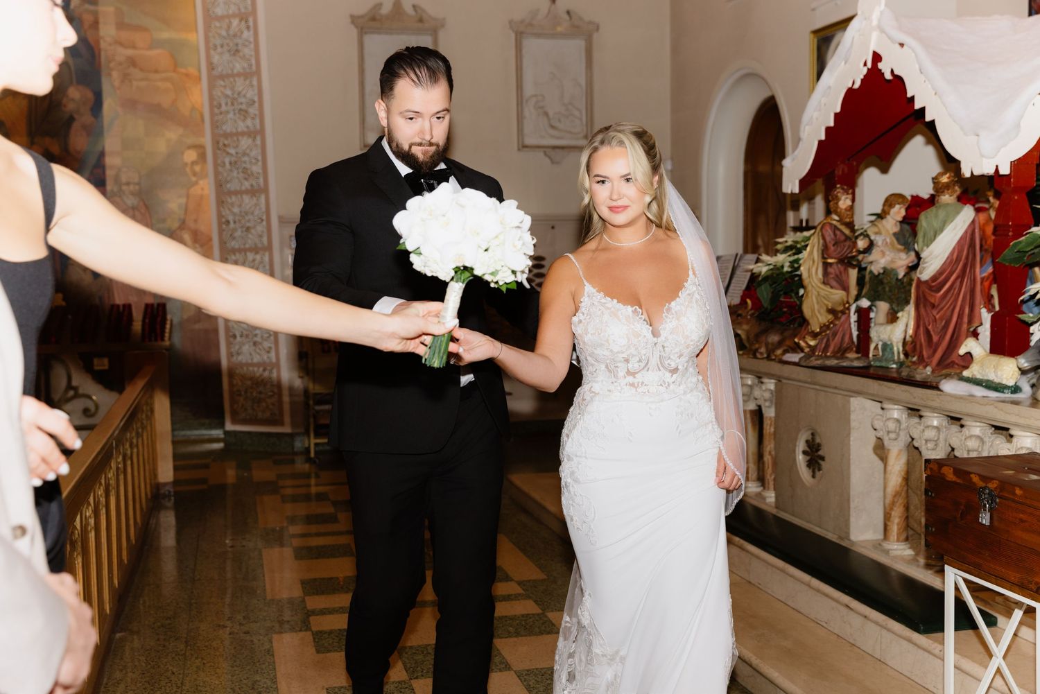 Bride in lace gown holding white bouquet walks with groom past ornate church decor and Christmas tree.