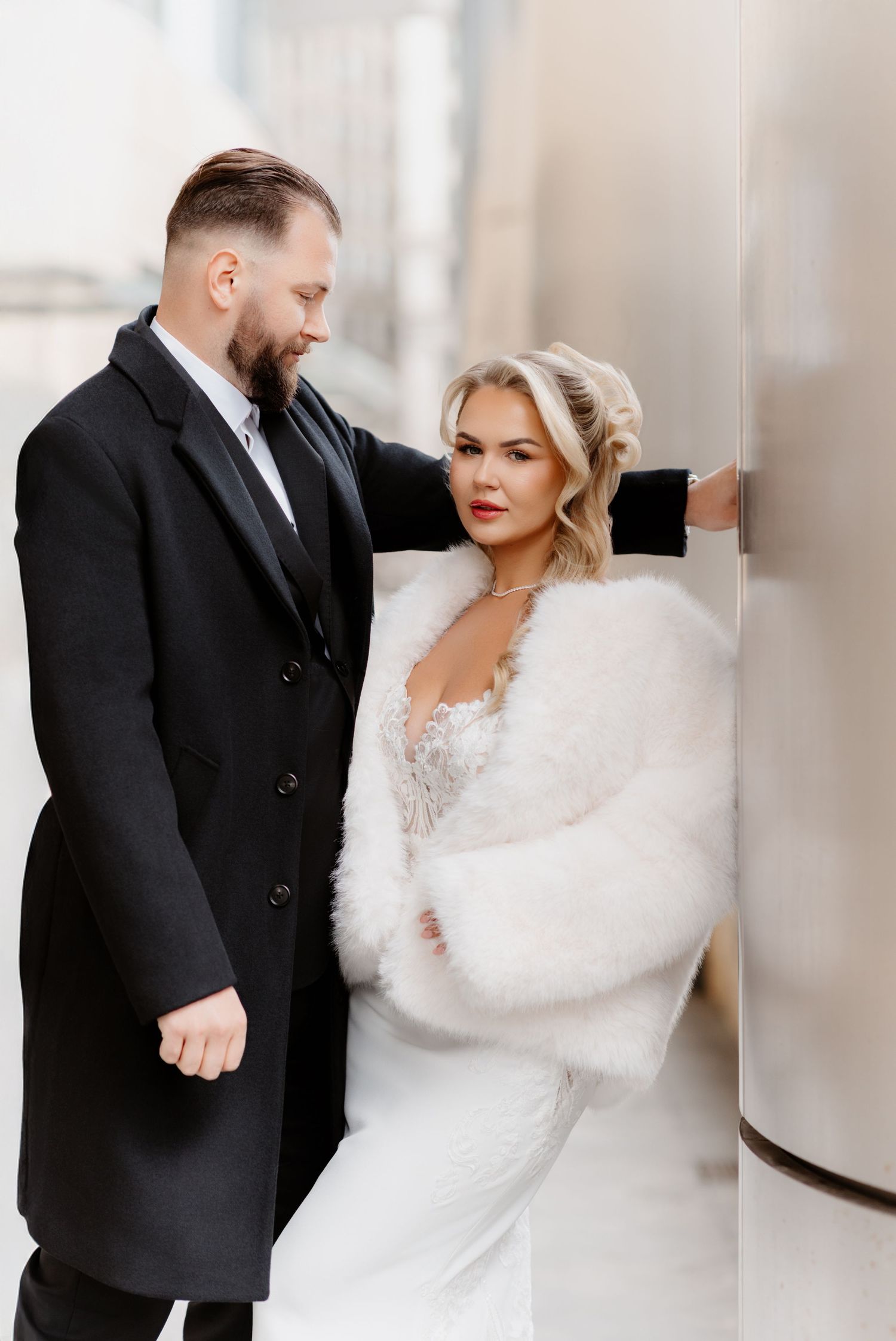 Wedding couple poses by metallic column, bride in white lace dress and fur stole, groom in black coat.