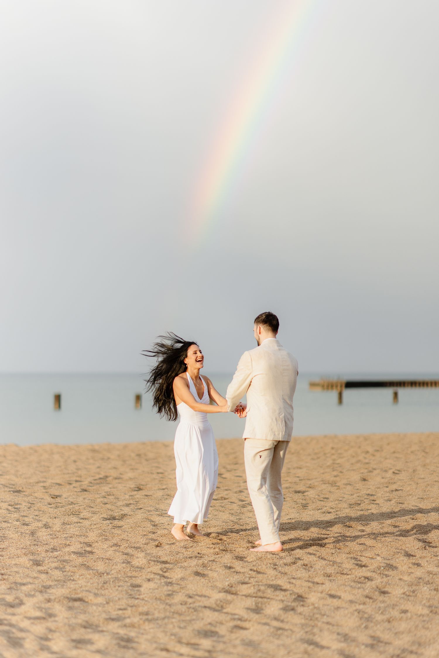 A happy couple holding hands and dancing on a beach under a beautiful rainbow in the sky.