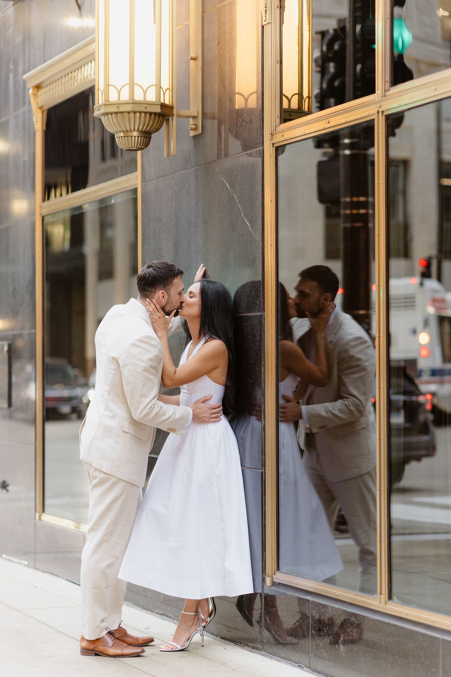 Couple kissing beside elegant gold-trimmed building windows reflected in glass on urban street.