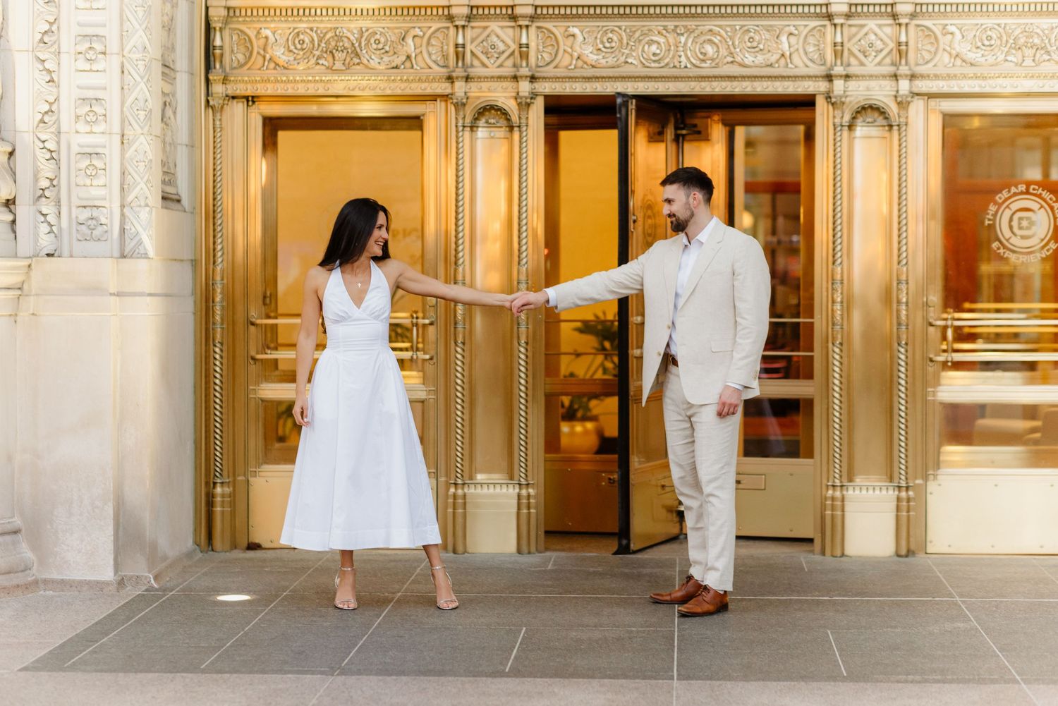 Couple in white and beige outfits holding hands outside a gold Art Deco building entrance in Chicago.