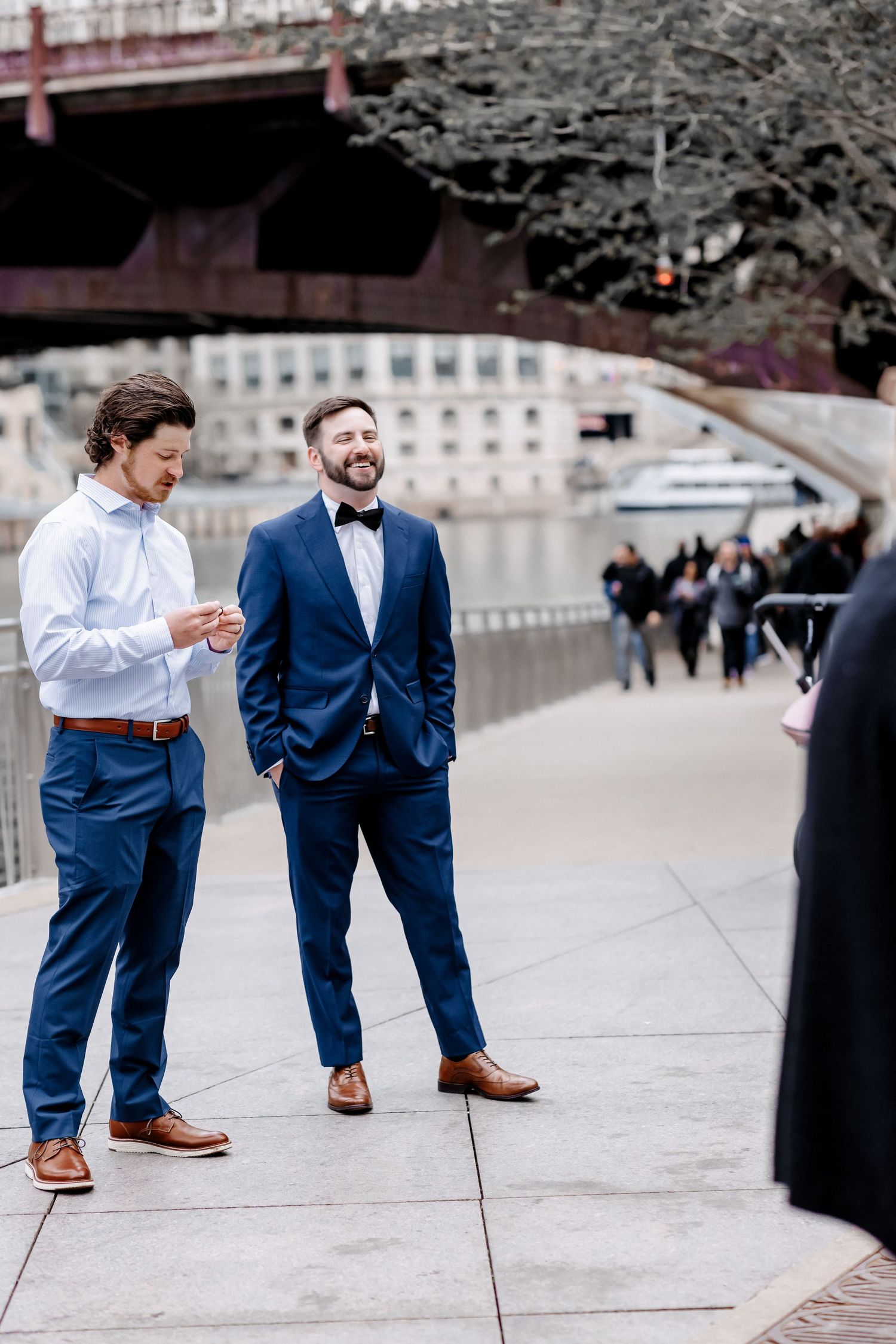 Two men in blue suits laugh together on a city riverwalk with a bridge in the background.