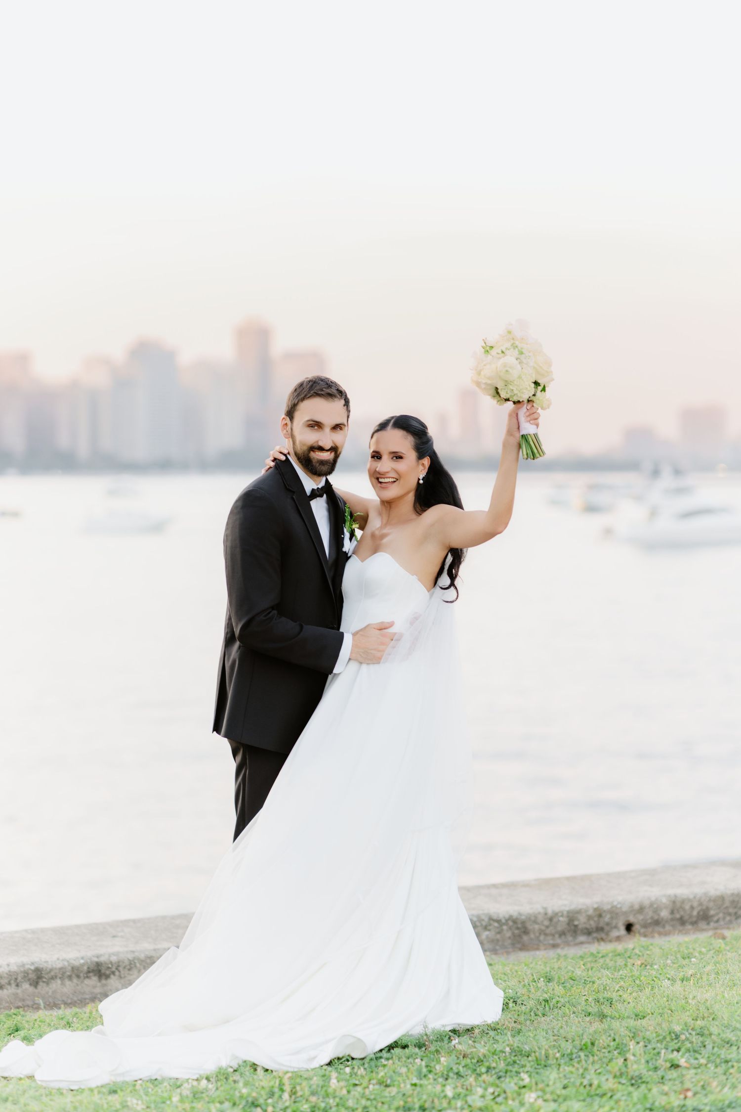 Joyful bride raises white bouquet while groom embraces her by the waterfront with a glowing city skyline.