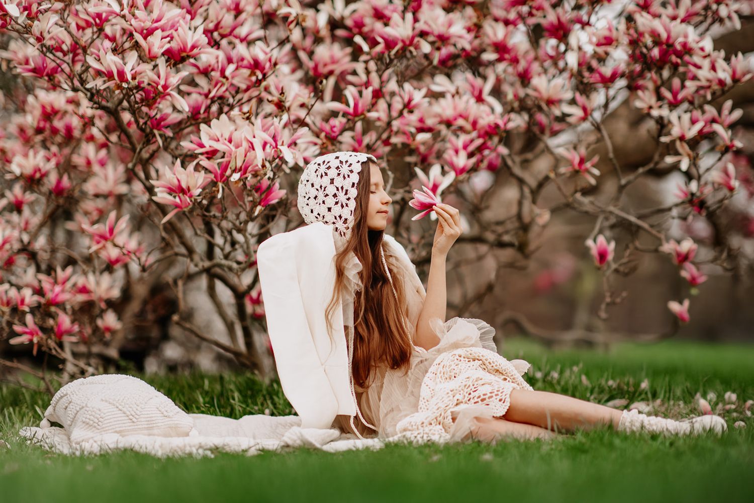 Young woman in lace dress sitting under blooming pink magnolia tree, holding a flower in spring garden.