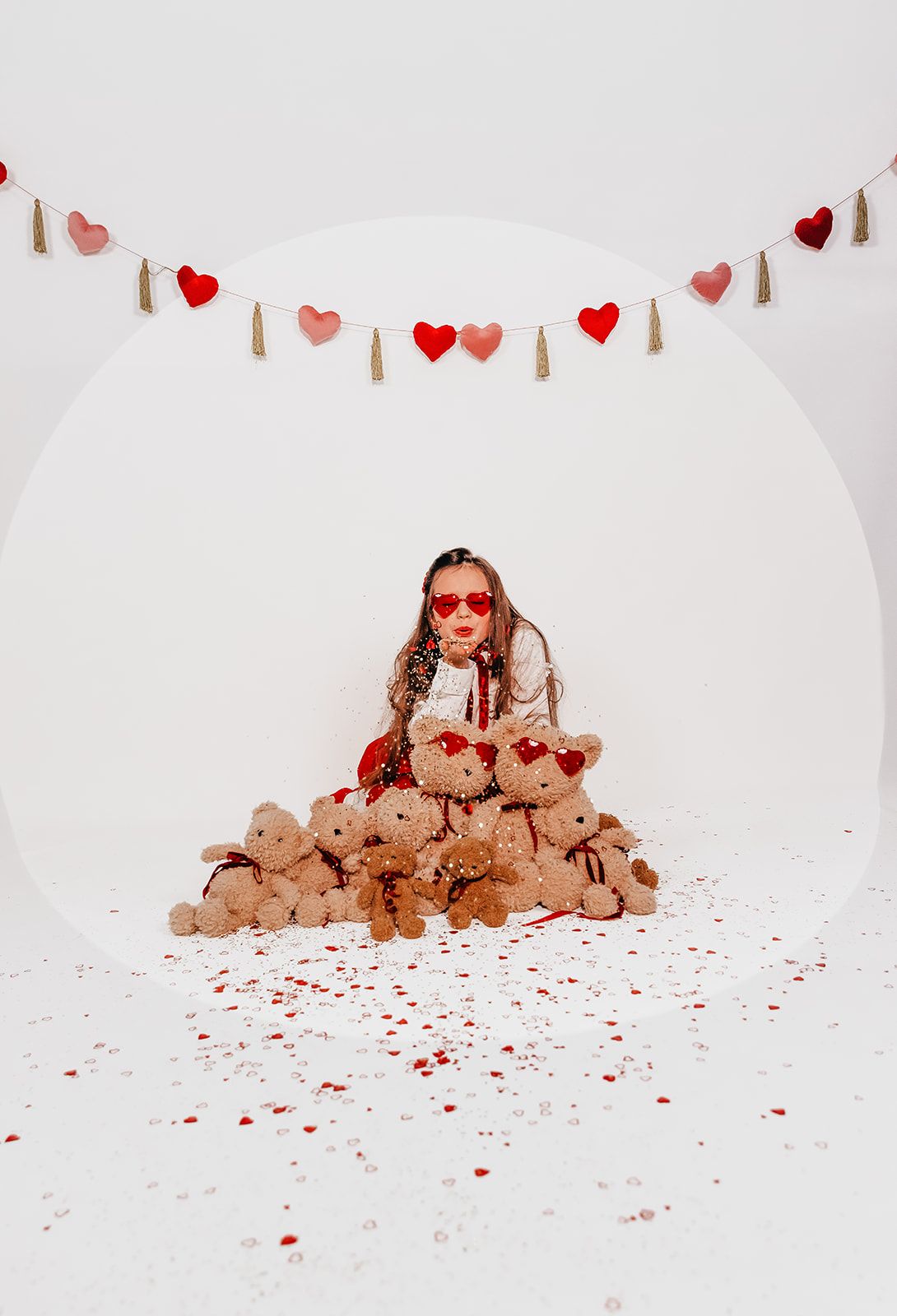 Girl in heart sunglasses sitting among teddy bears with Valentine's Day heart garland and confetti on white background.