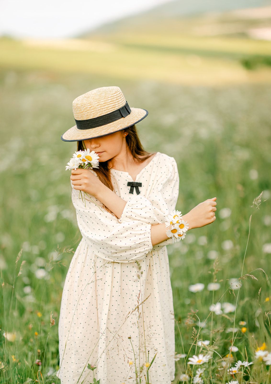 Young woman in polka dot dress and straw hat smelling white daisies in a blooming meadow field.