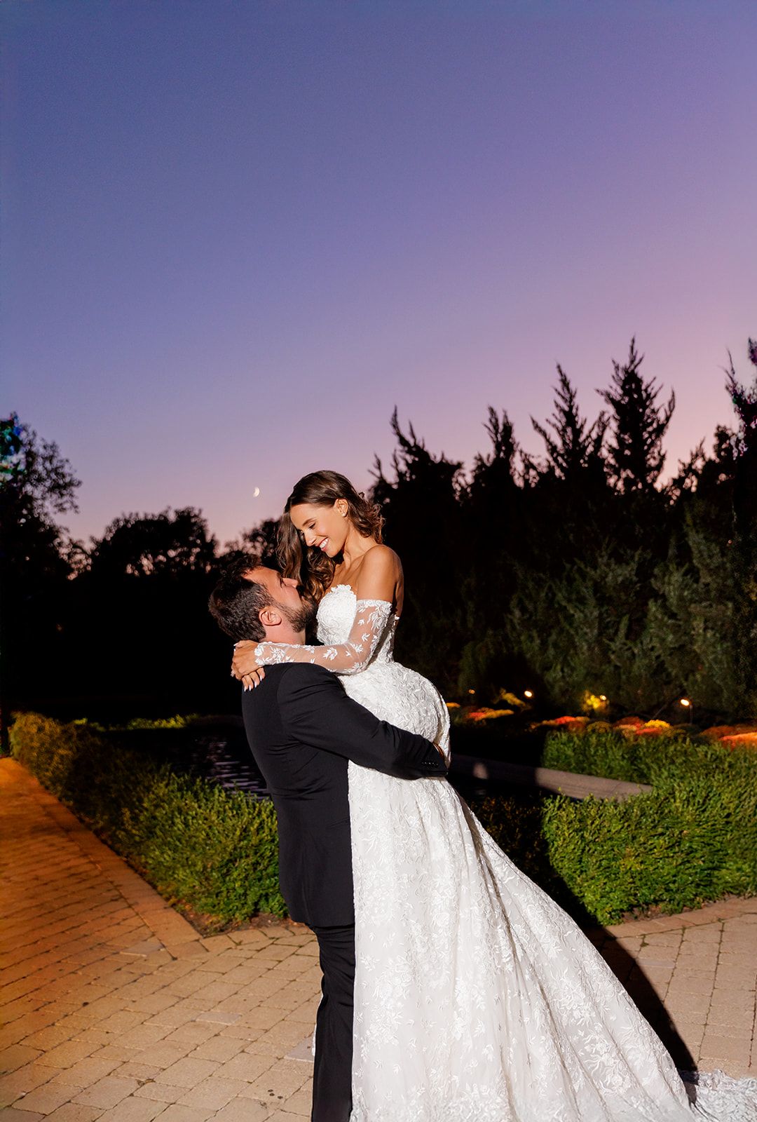 Groom lifts bride joyfully as she smiles down at him under a romantic purple twilight sky.