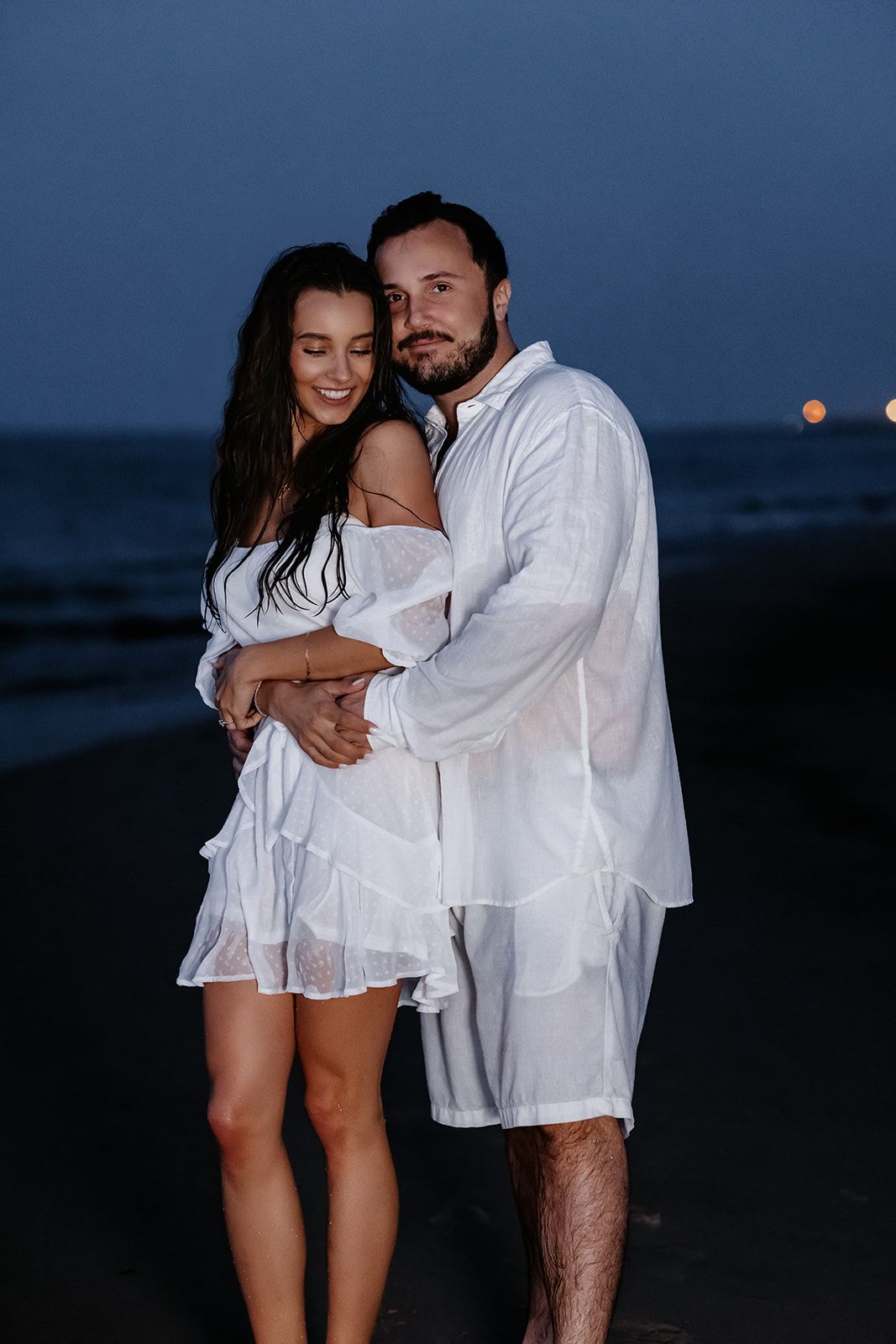 Romantic couple in white outfits embracing on a beach at dusk with ocean waves in the background.