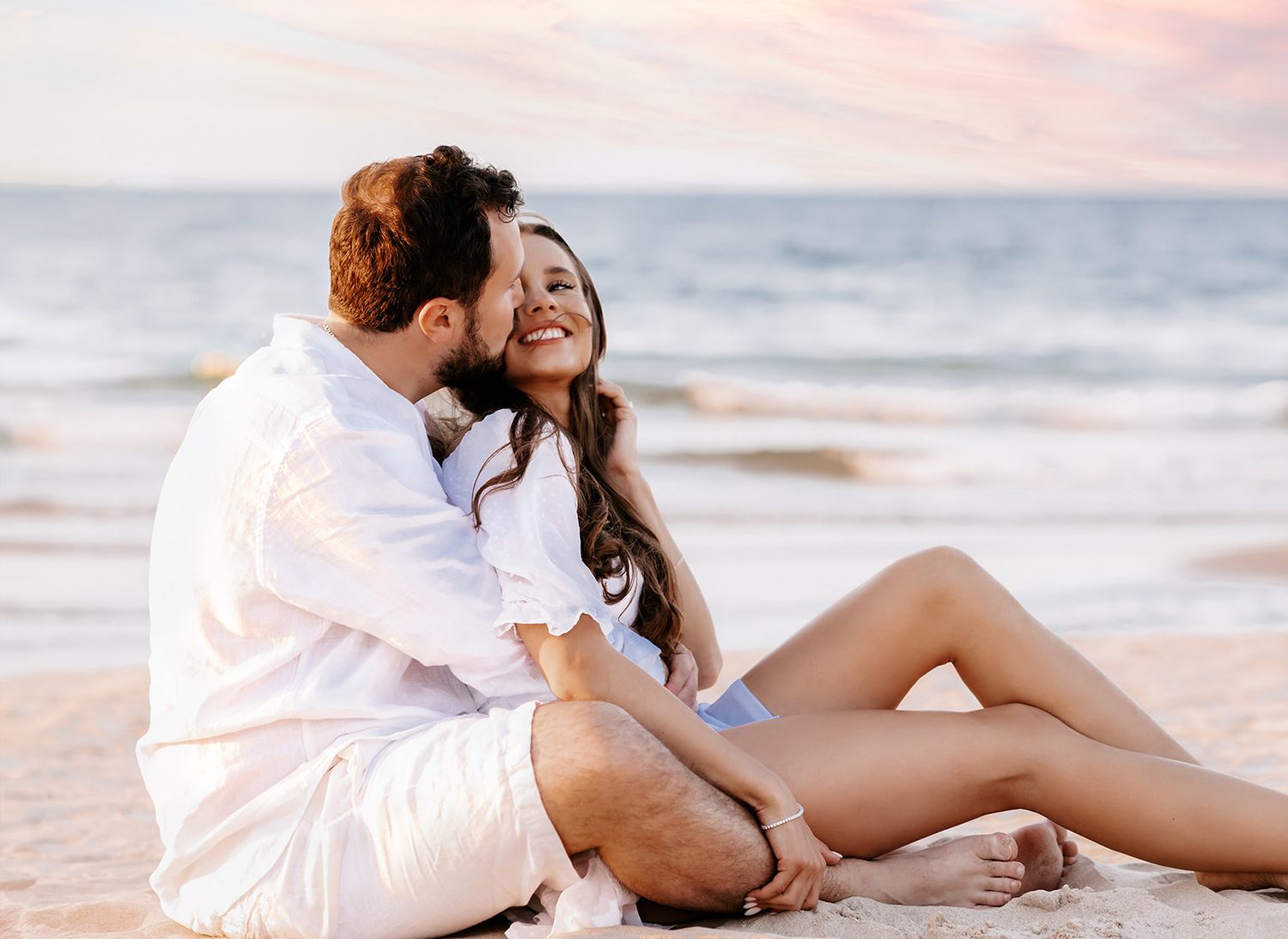 Couple sitting on beach sand at sunset, man nuzzling smiling woman affectionately.
