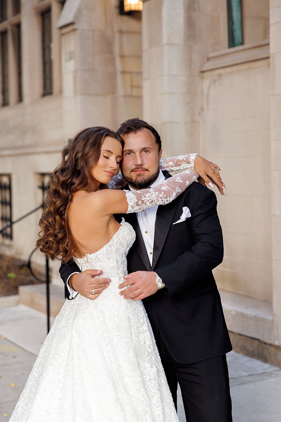 Bride in backless lace gown embraces groom in black tuxedo outdoors near stone building.