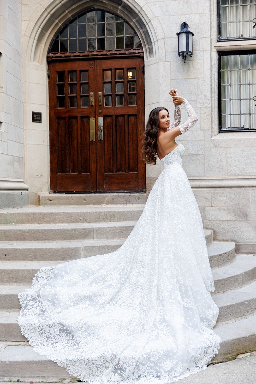 Bride in elegant white lace gown with long train poses on stone steps before ornate wooden doors.