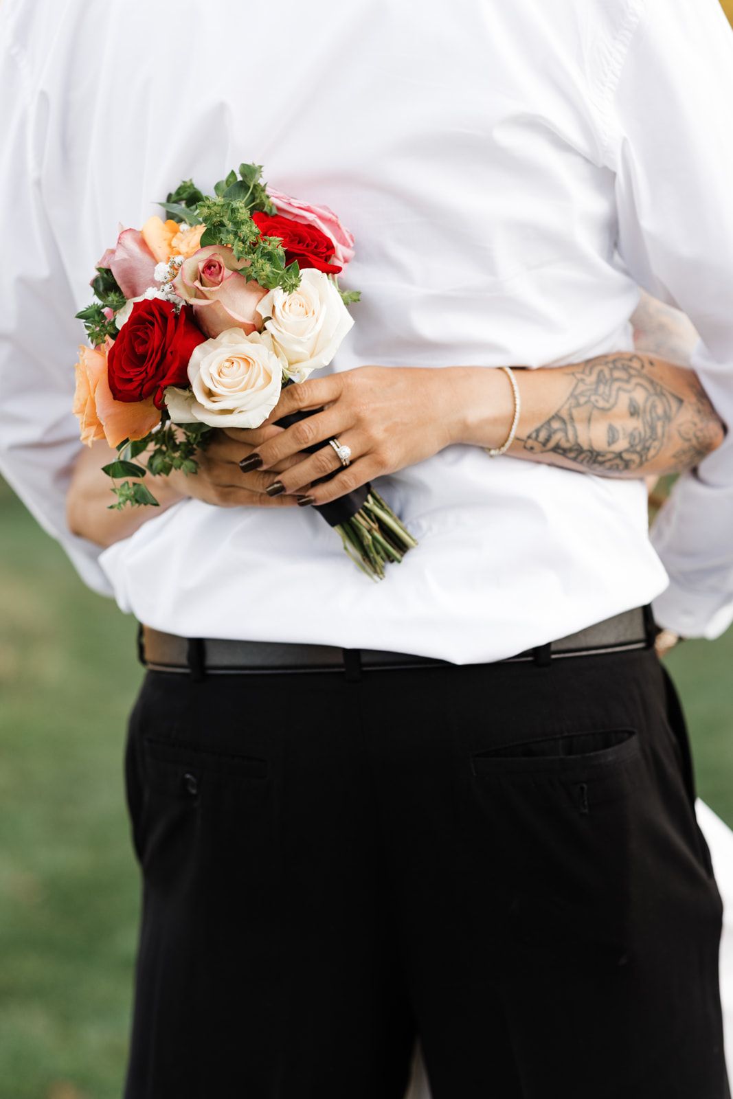 Bride with tattooed arm holding colorful rose bouquet behind groom in white shirt and black pants.