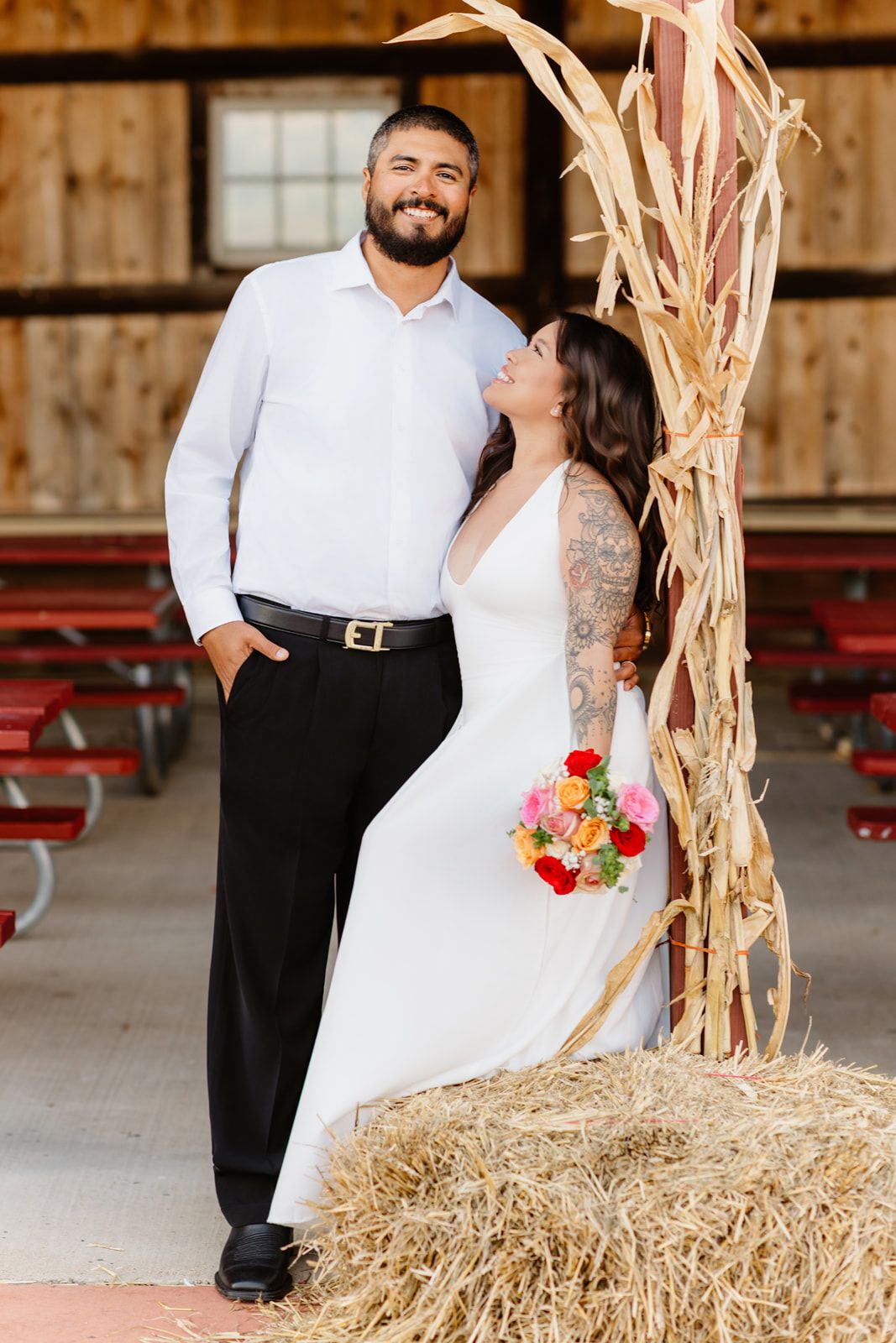 A bride and groom pose together on hay bales at a rustic barn wedding venue.