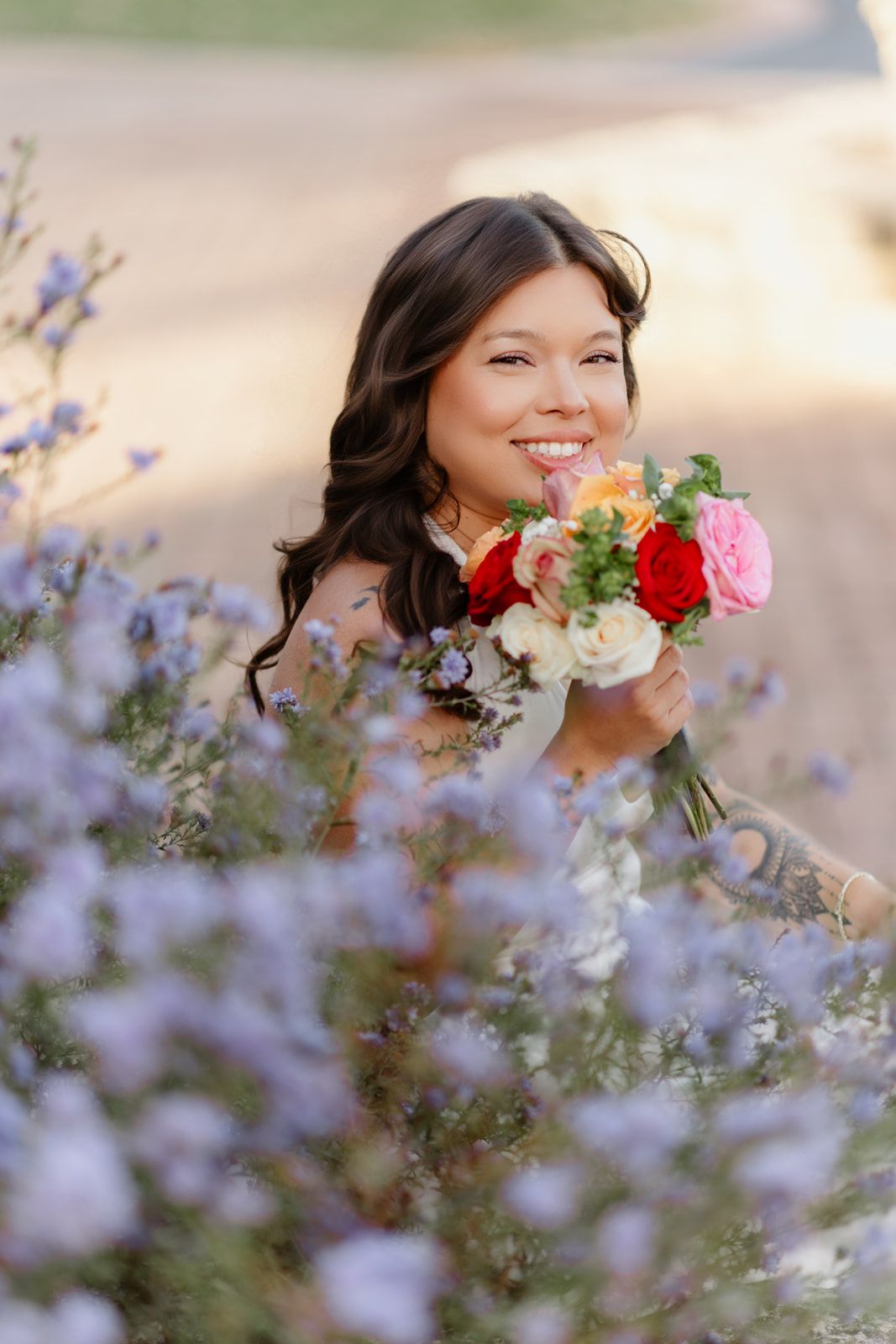 Smiling bride holds a colorful bouquet of red, pink, and white roses while sitting among purple wildflowers.