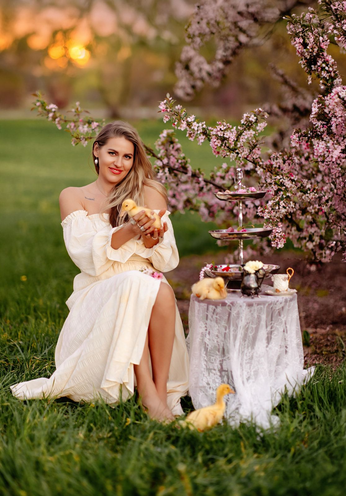 Woman in white off-shoulder dress holding ducklings under pink cherry blossoms at a spring garden tea party.