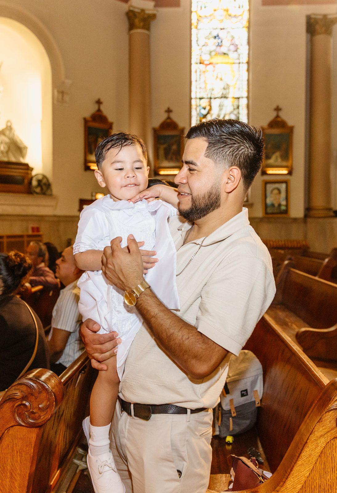 A man holds a young child in a white outfit inside a church with wooden pews and stained glass windows.
