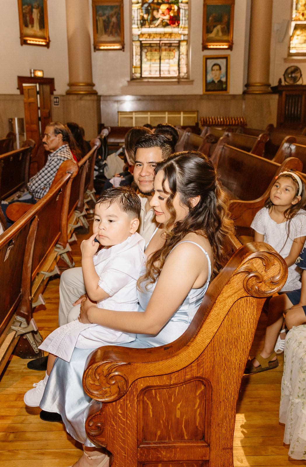 Young family with toddler boy in white outfit sitting in wooden church pews during baptism mass.
