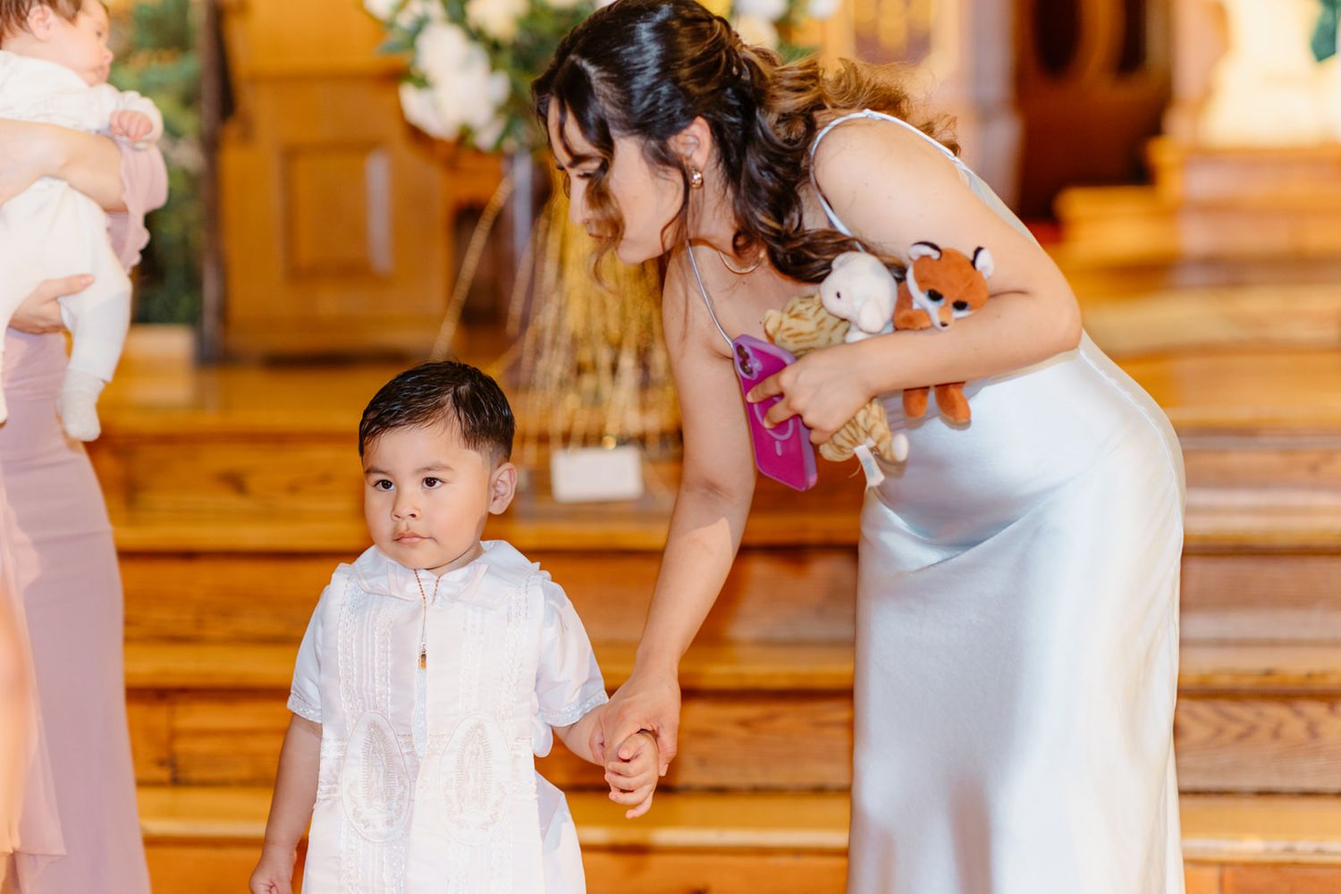 Woman in silver dress kneels to hold hands with young boy in white outfit inside a wooden church.