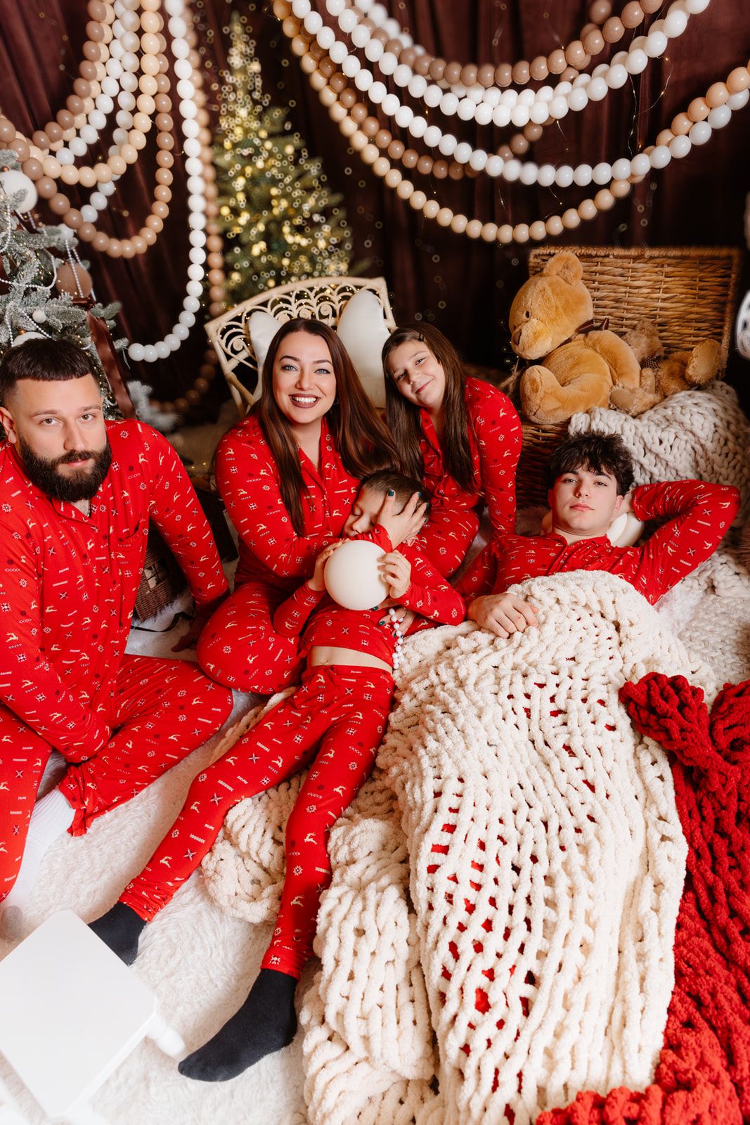 Family of four in matching red Christmas pajamas relaxing together on a cozy bed with holiday decorations.