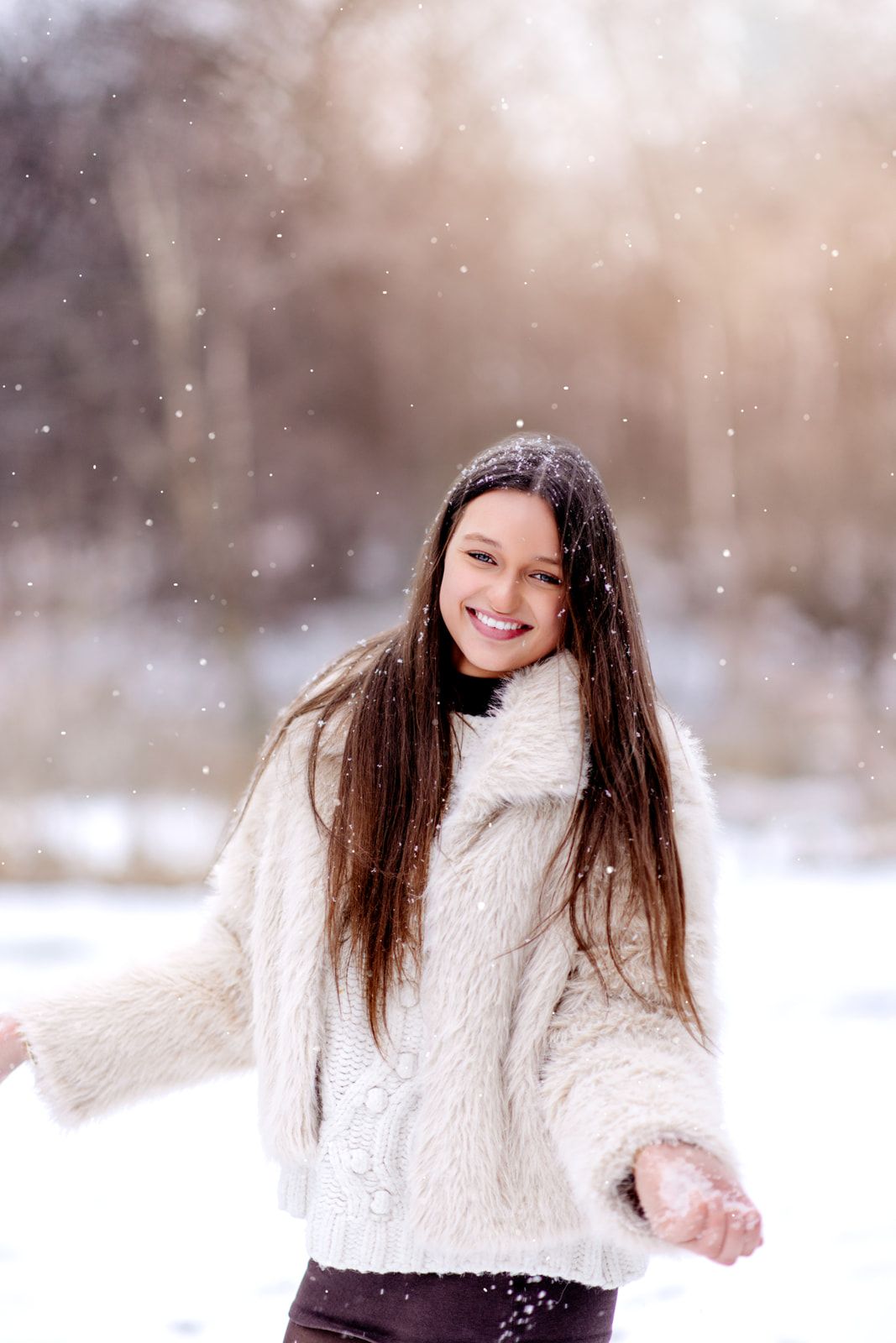 A joyful teenage girl in a cream faux fur coat laughs and plays in falling snow in a bright winter landscape.