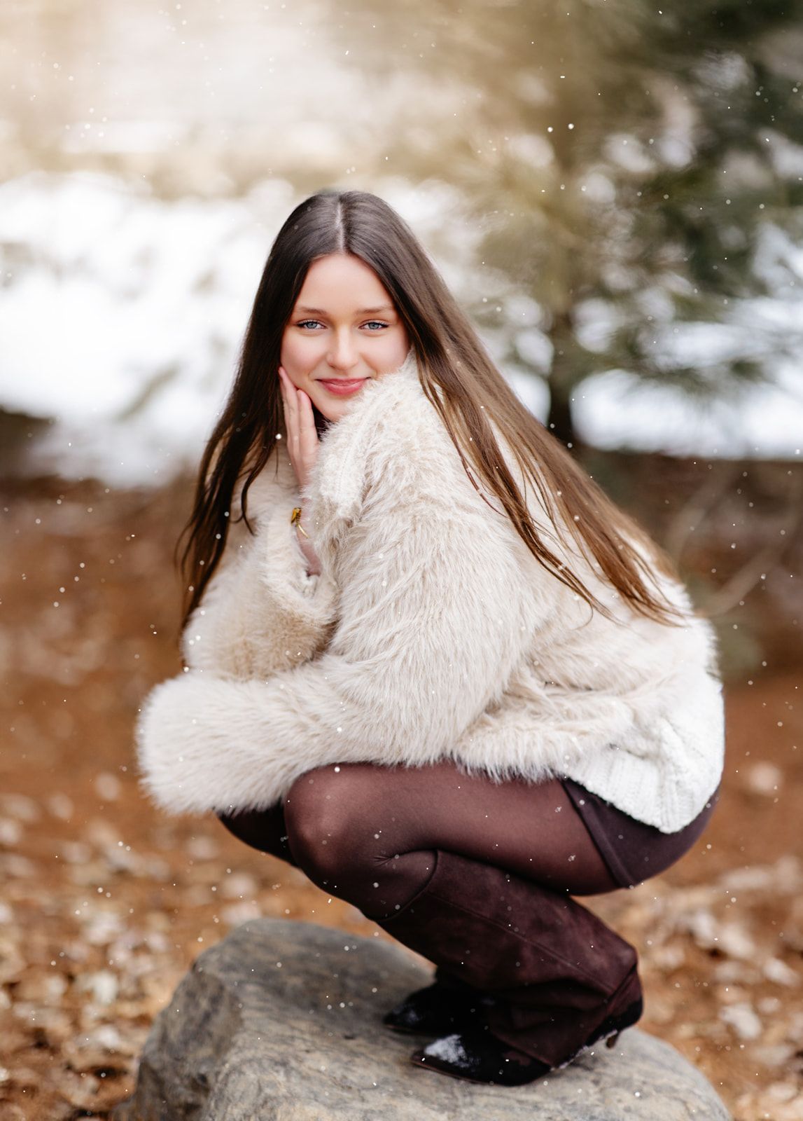Young woman in fluffy cream sweater crouching on a rock in a snowy winter forest setting.