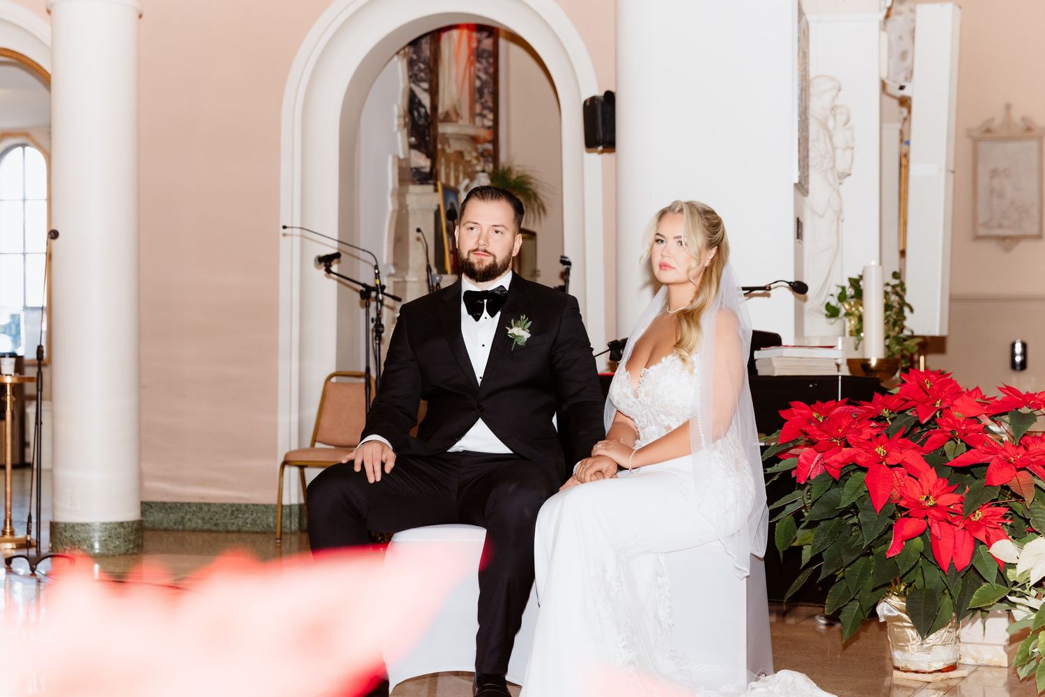 Bride and groom sitting at church altar during ceremony with vibrant red poinsettia arrangements.
