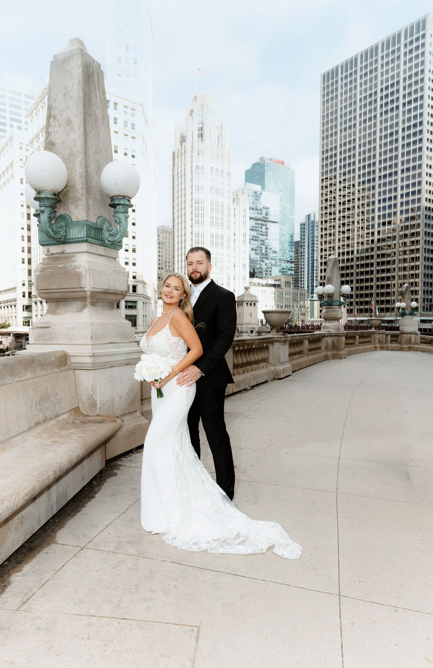 Bride in fitted lace gown and groom pose on Chicago Riverwalk bridge with city skyline.