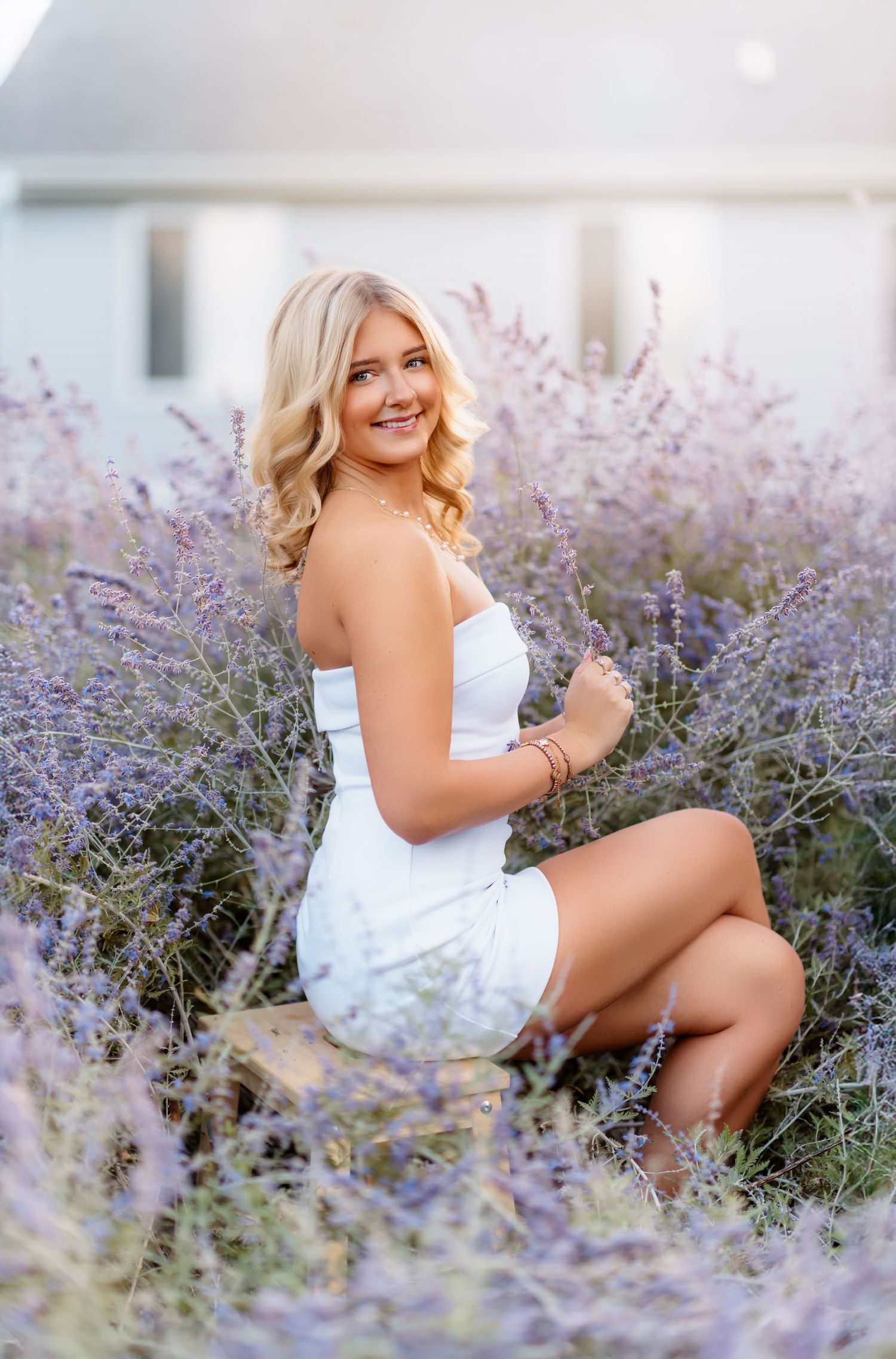 Young blonde woman in white dress sitting among purple lavender flowers, smiling over shoulder.