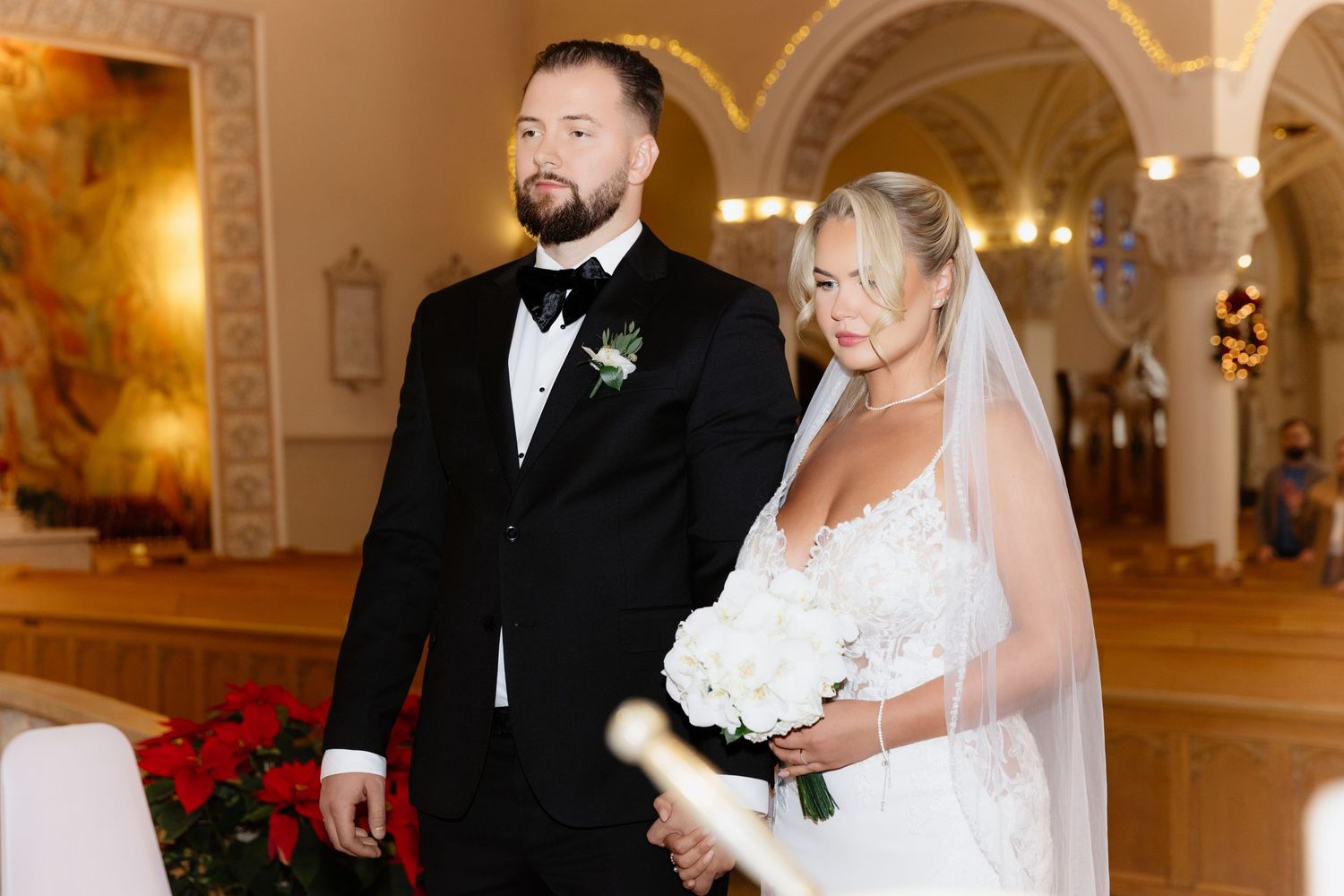 Bride in white gown holding bouquet stands beside groom in black tuxedo during church wedding ceremony.