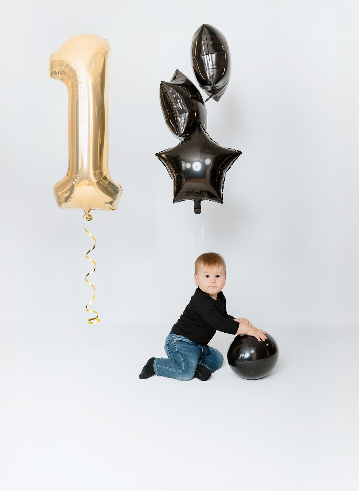 Baby boy in black outfit sits beside gold number one and black star balloons for first birthday photos.