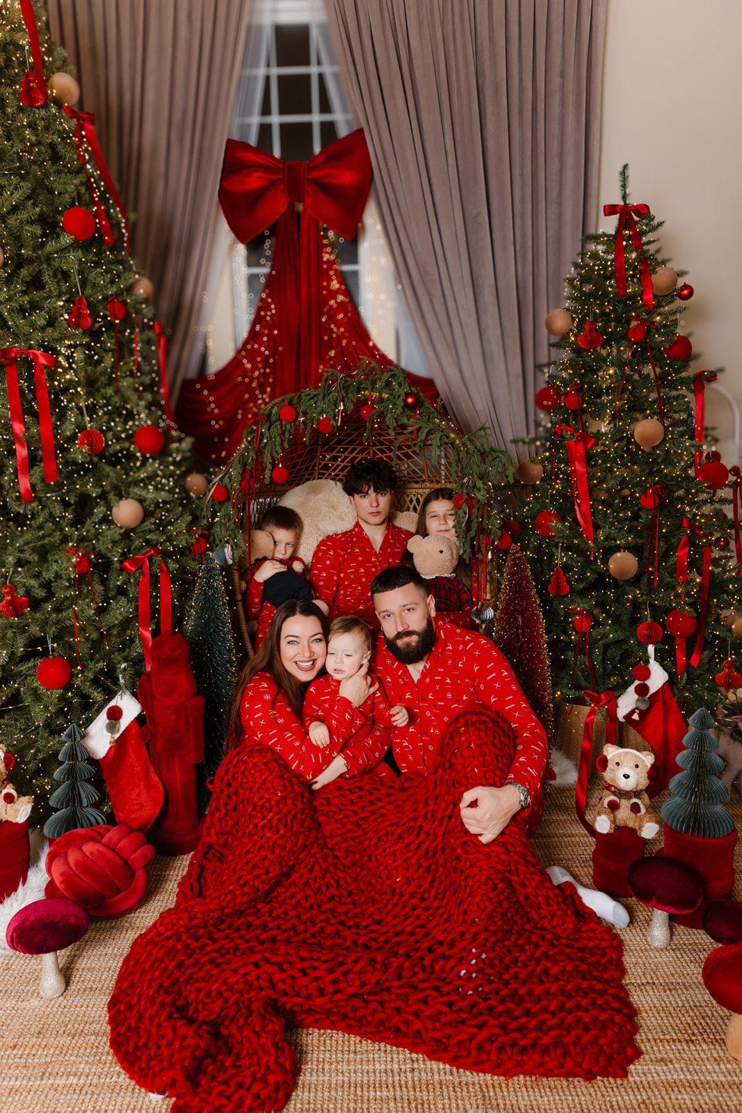 Family in matching red pajamas poses by decorated Christmas trees with red bows and ornaments during holiday celebration.