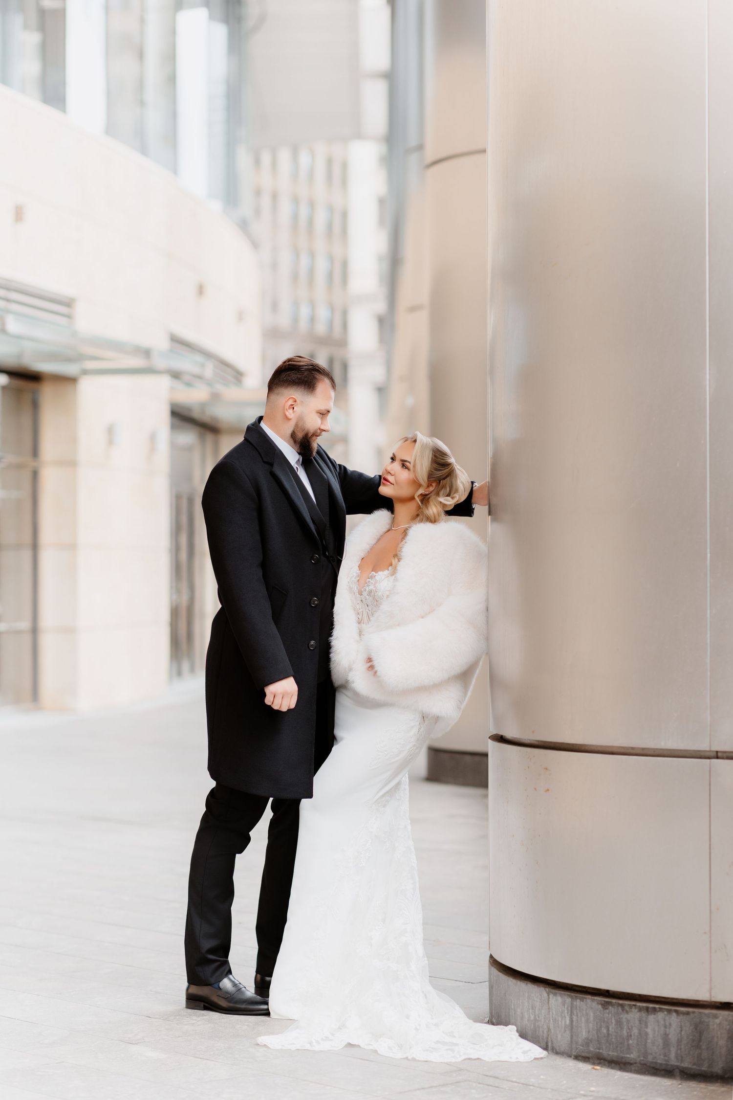 Bride in white lace gown with fur wrap and groom in black coat posing by urban column.