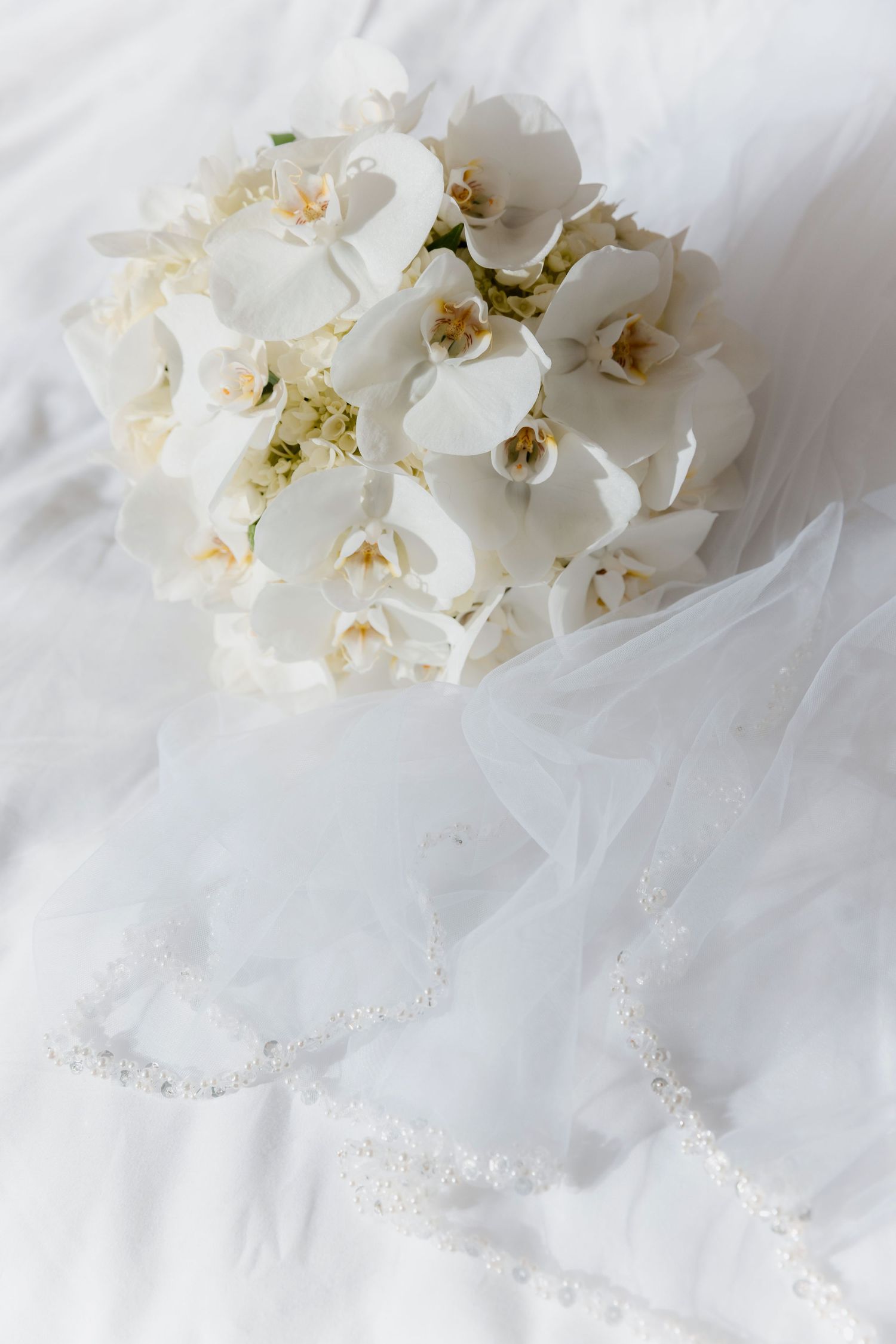 White orchid bridal bouquet resting on white veil with pearl necklace on soft white fabric.