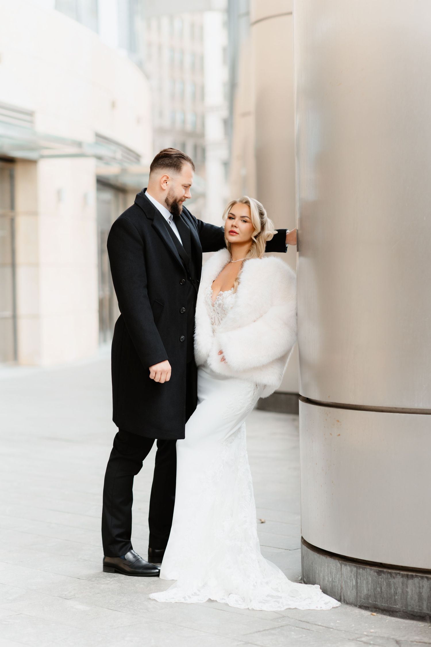 Bride in white fur wrap and groom in black coat pose by silver columns in urban setting.