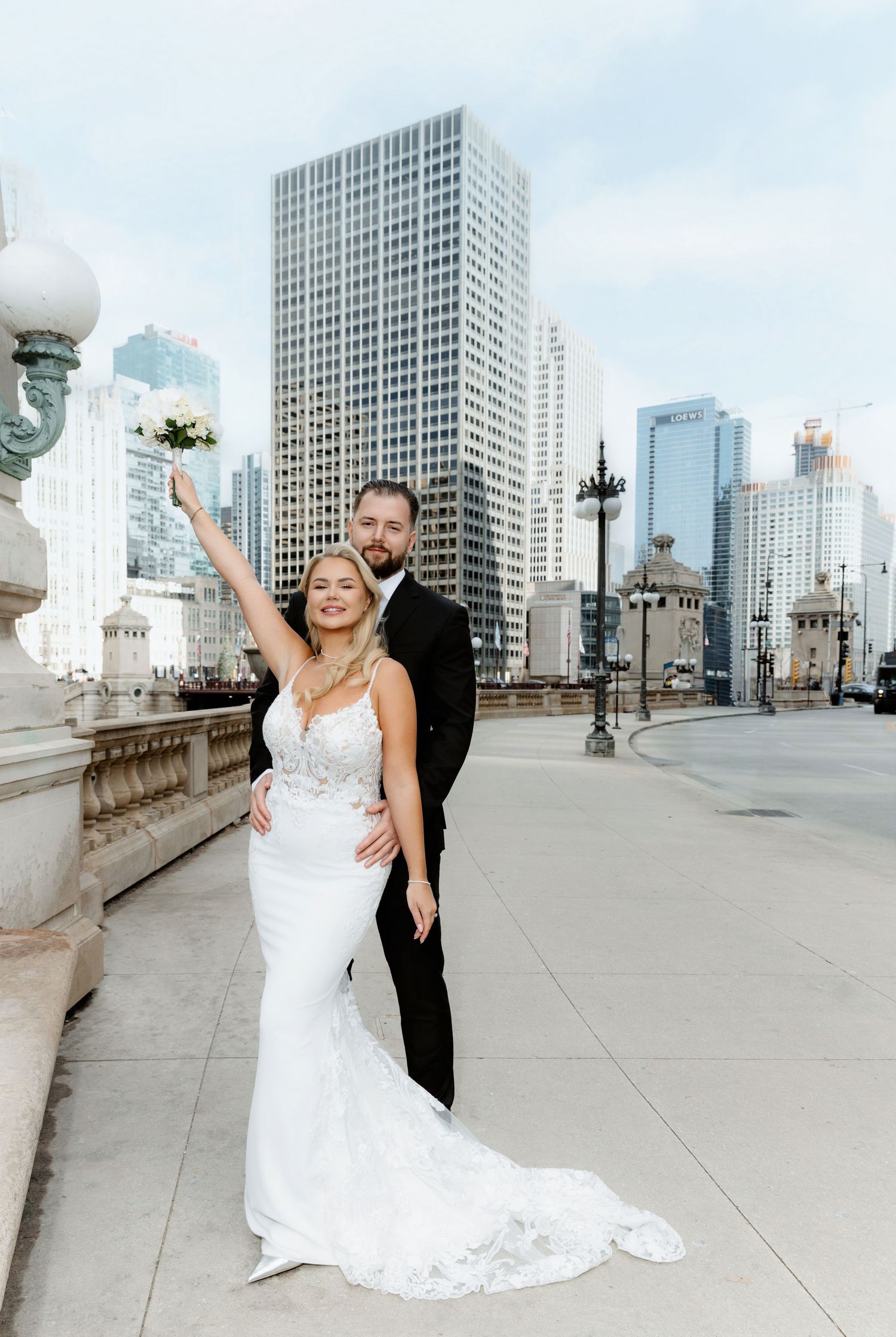 Bride in lace gown raises bouquet while groom in tuxedo embraces her, posing on Chicago city bridge.