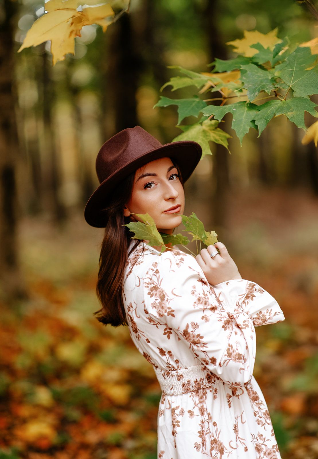 Young woman in floral dress and brown hat holding maple leaves in an autumn forest.