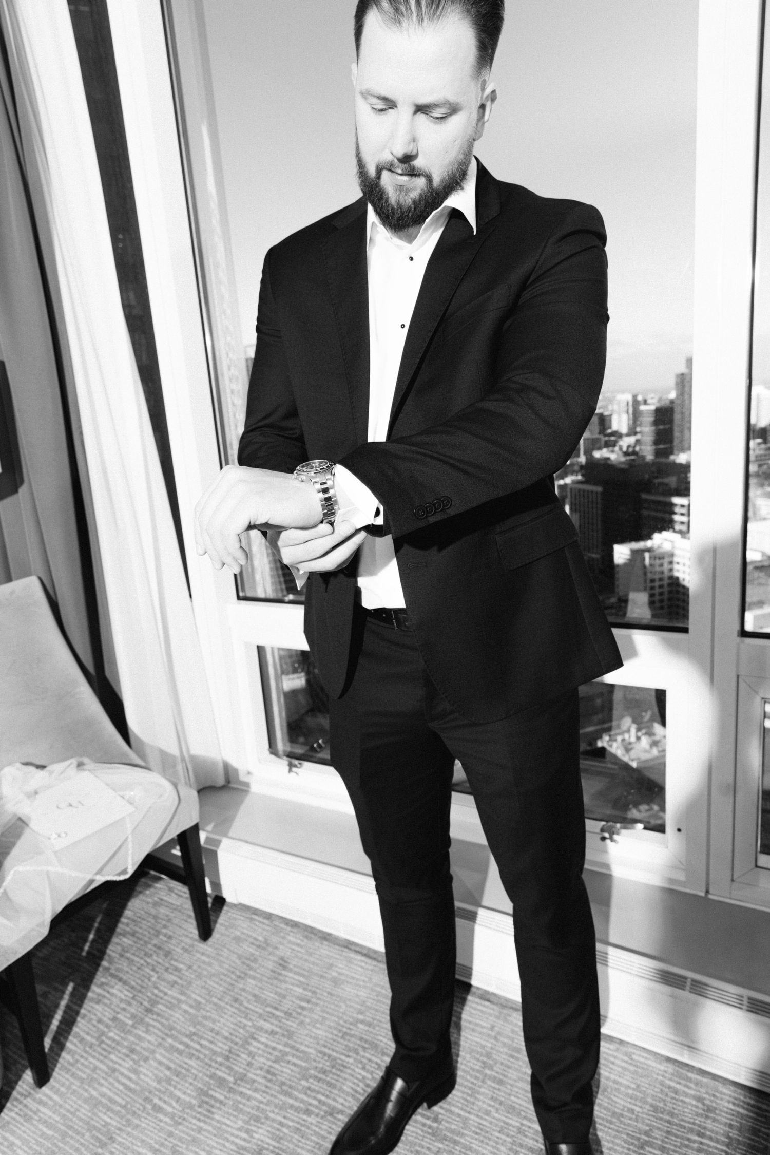 Black and white photo of bearded groom in black suit adjusting cufflinks by a high-rise window with city view.