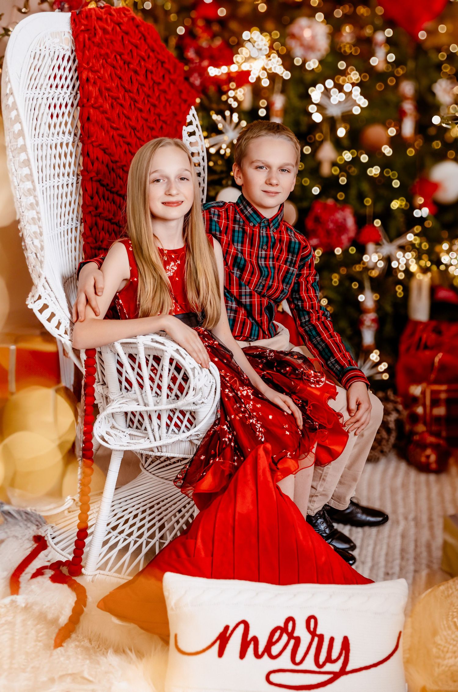 Two siblings in festive outfits pose together in white wicker chair near Christmas tree.