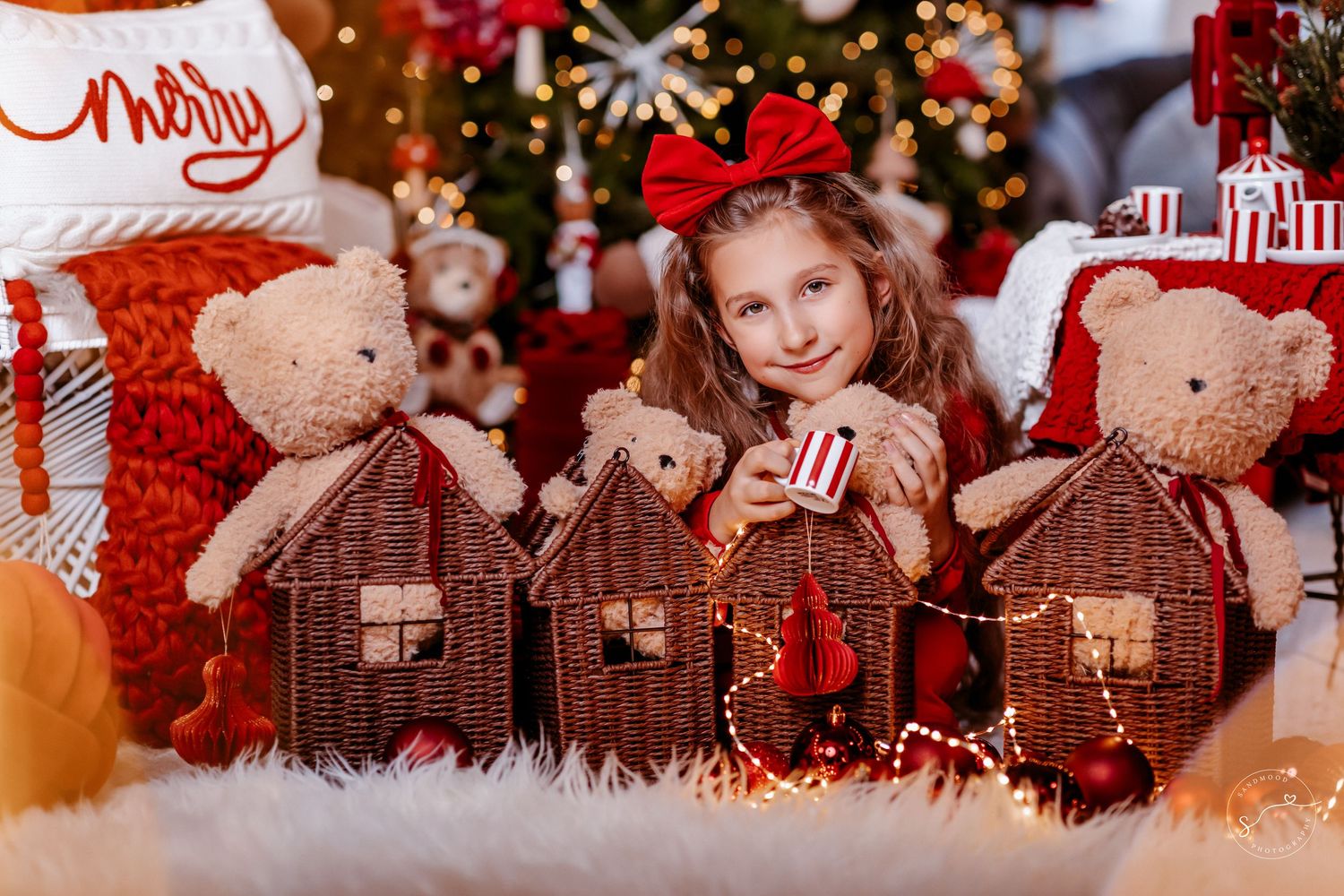 A smiling girl with a red bow headband poses with teddy bears and wicker houses near a Christmas tree.