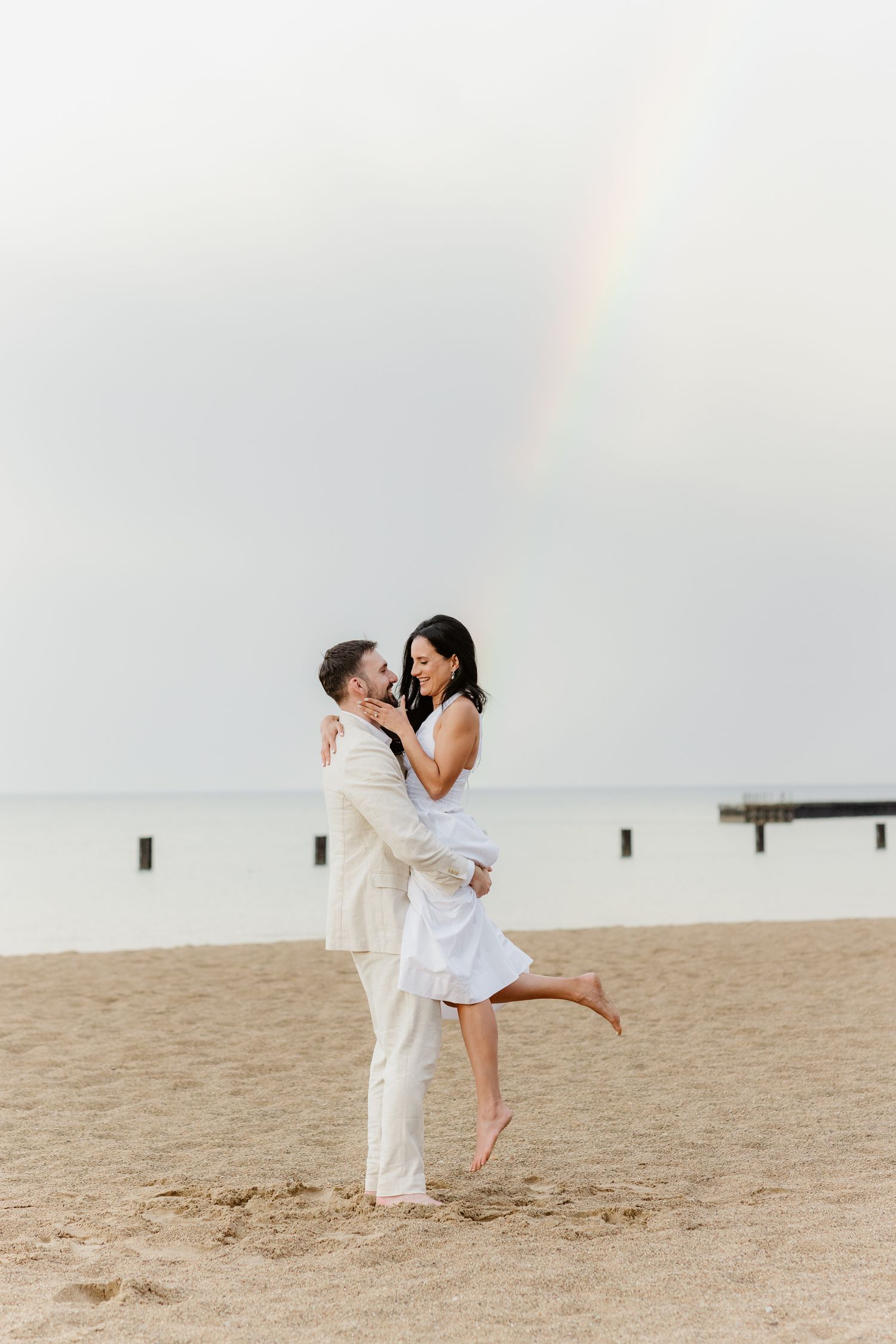 A couple embracing and smiling at each other on a beach with a rainbow over the ocean behind them.