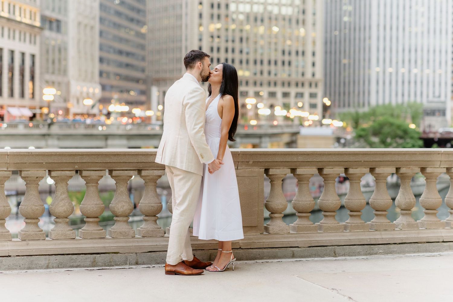 Couple standing close together on Chicago riverwalk balustrade, man in beige suit, woman in white midi dress.