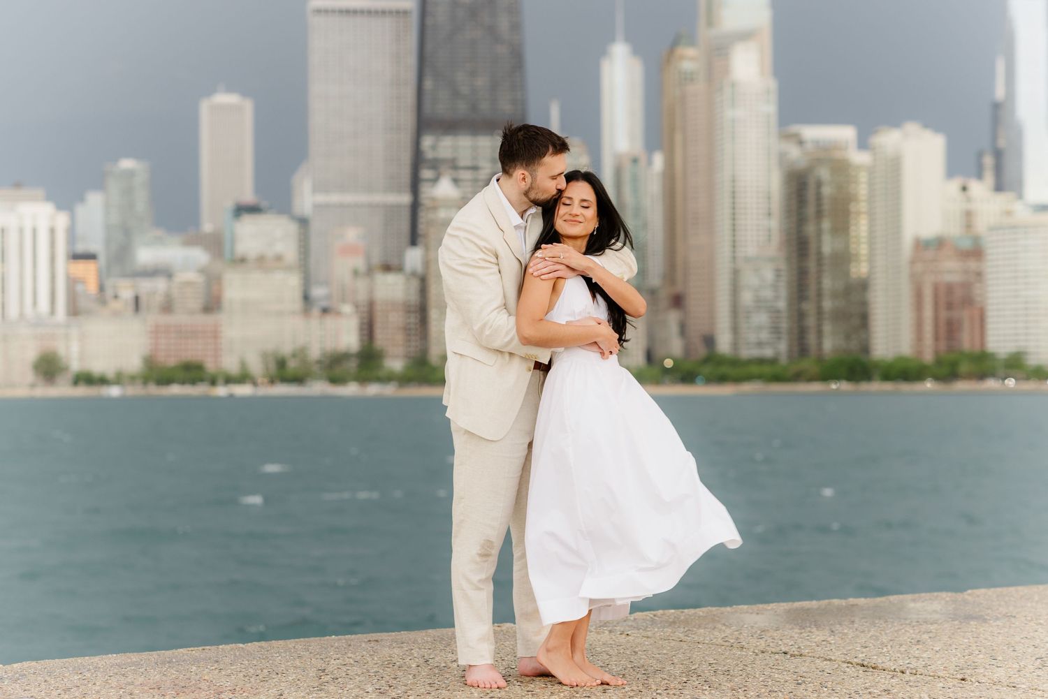 Couple embracing barefoot on Chicago lakefront with dramatic skyline and stormy sky in the background.