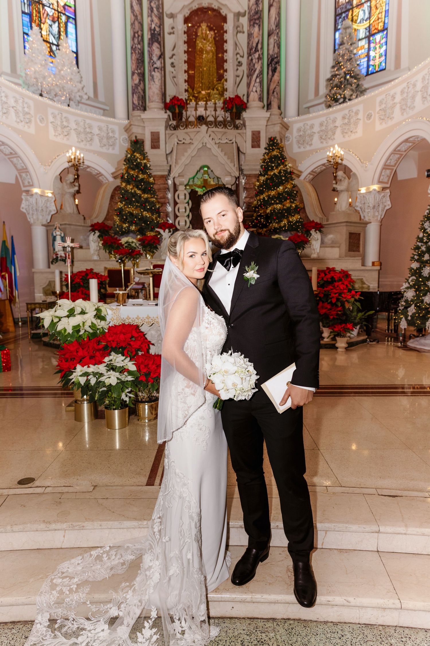 Bride and groom posing in decorated Christmas church with poinsettias and Christmas trees in background.