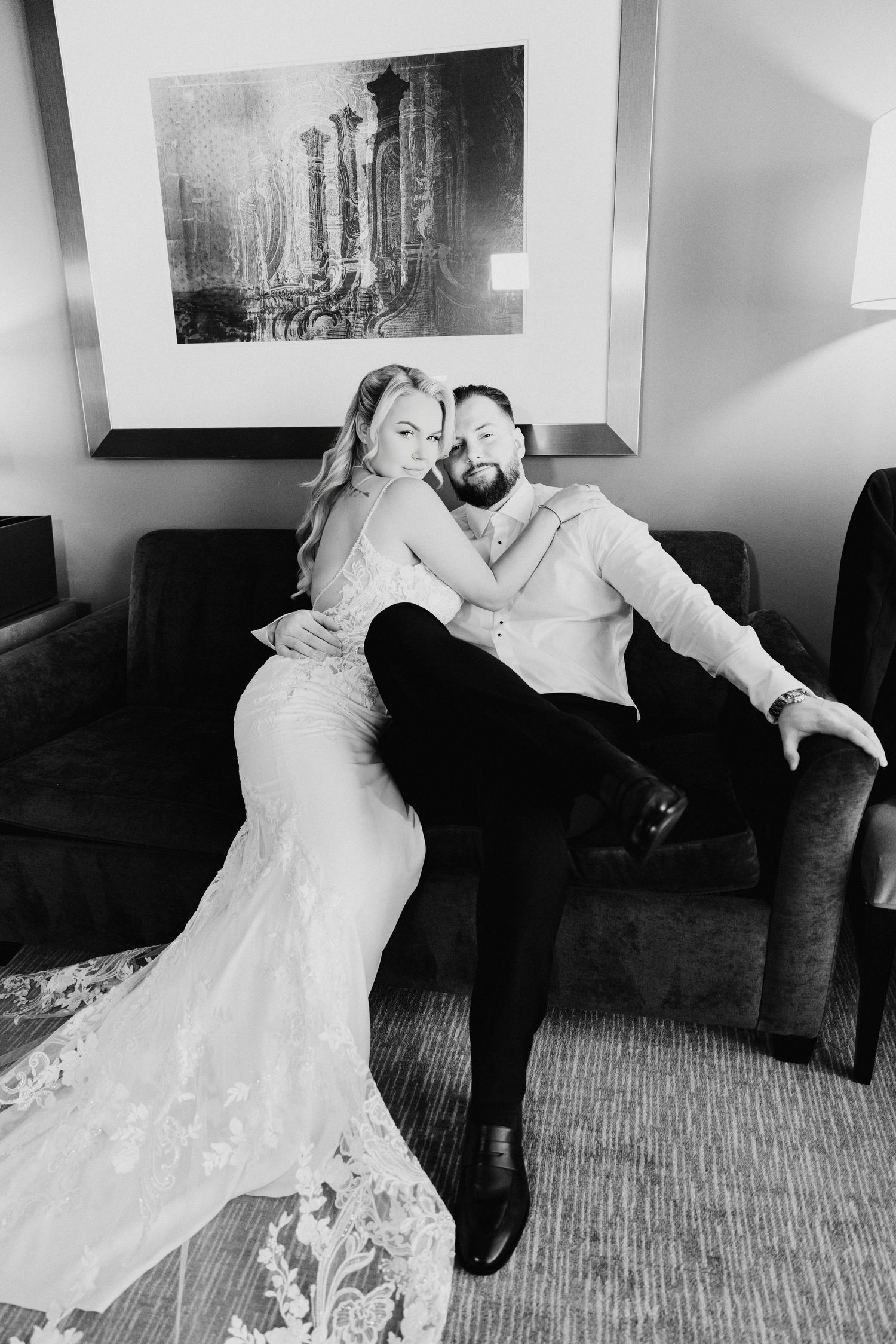 Bride in lace gown and groom embrace on sofa in black and white wedding portrait.
