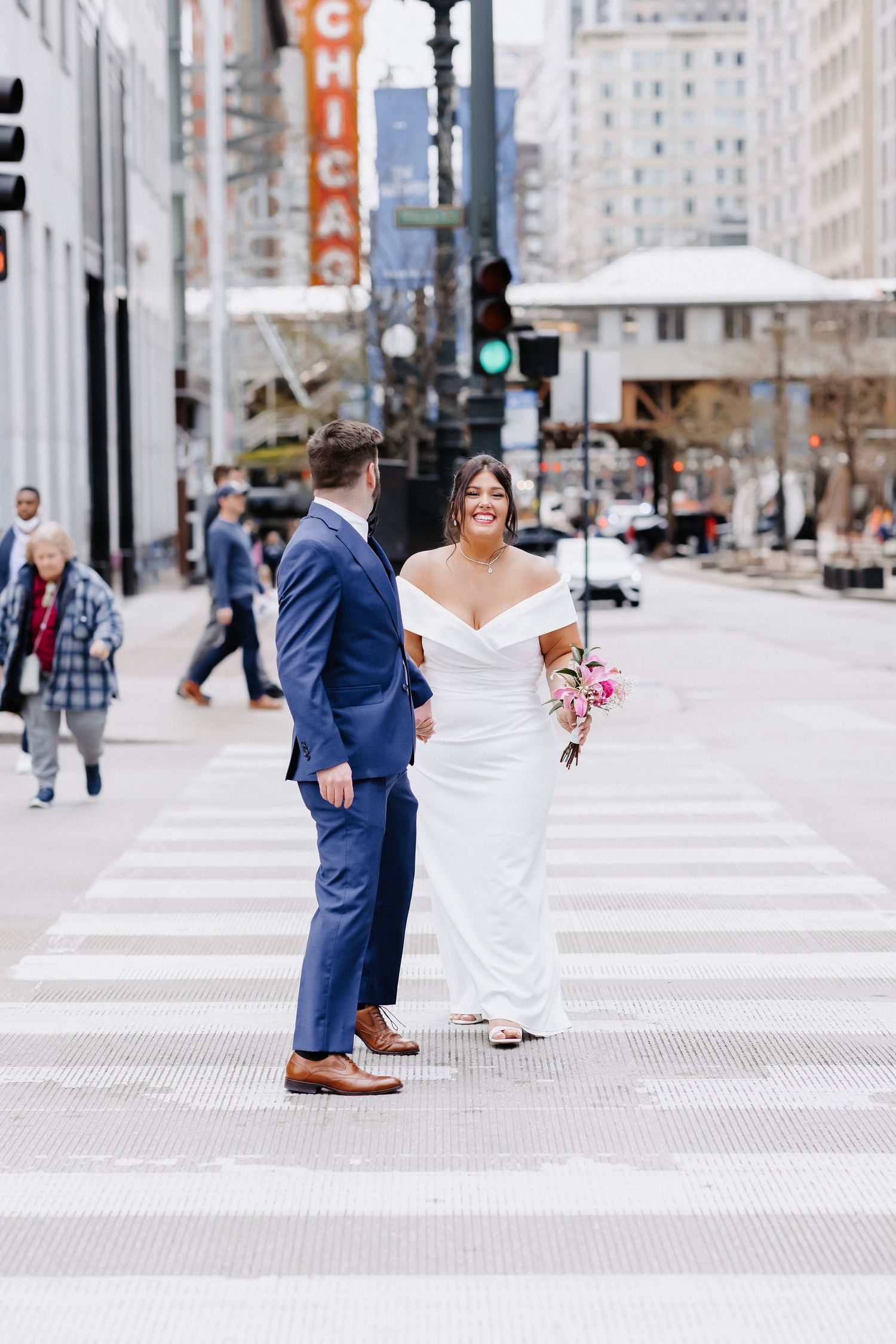Laughing bride in off-shoulder white gown and groom in navy suit cross Chicago street near Theatre marquee.